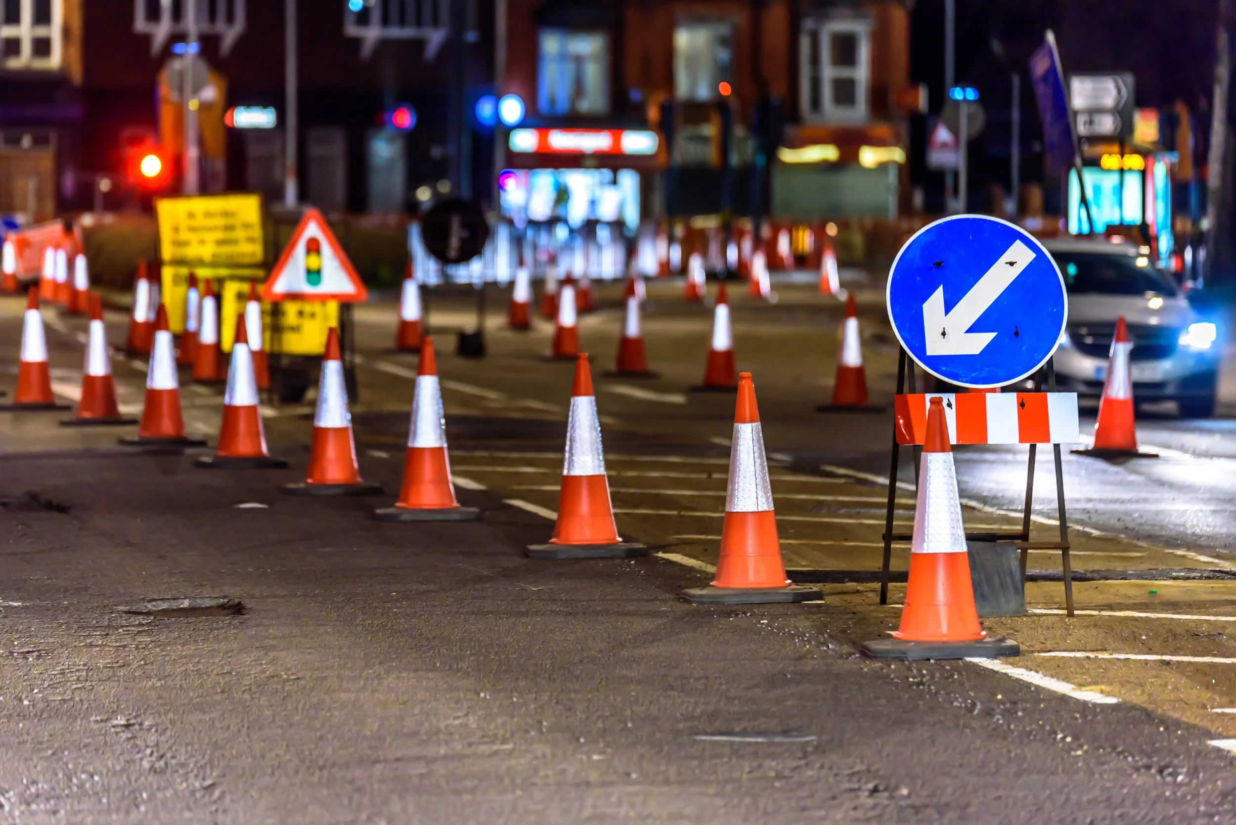 Nighttime construction zone with orange traffic cones and a blue directional arrow sign pointing left