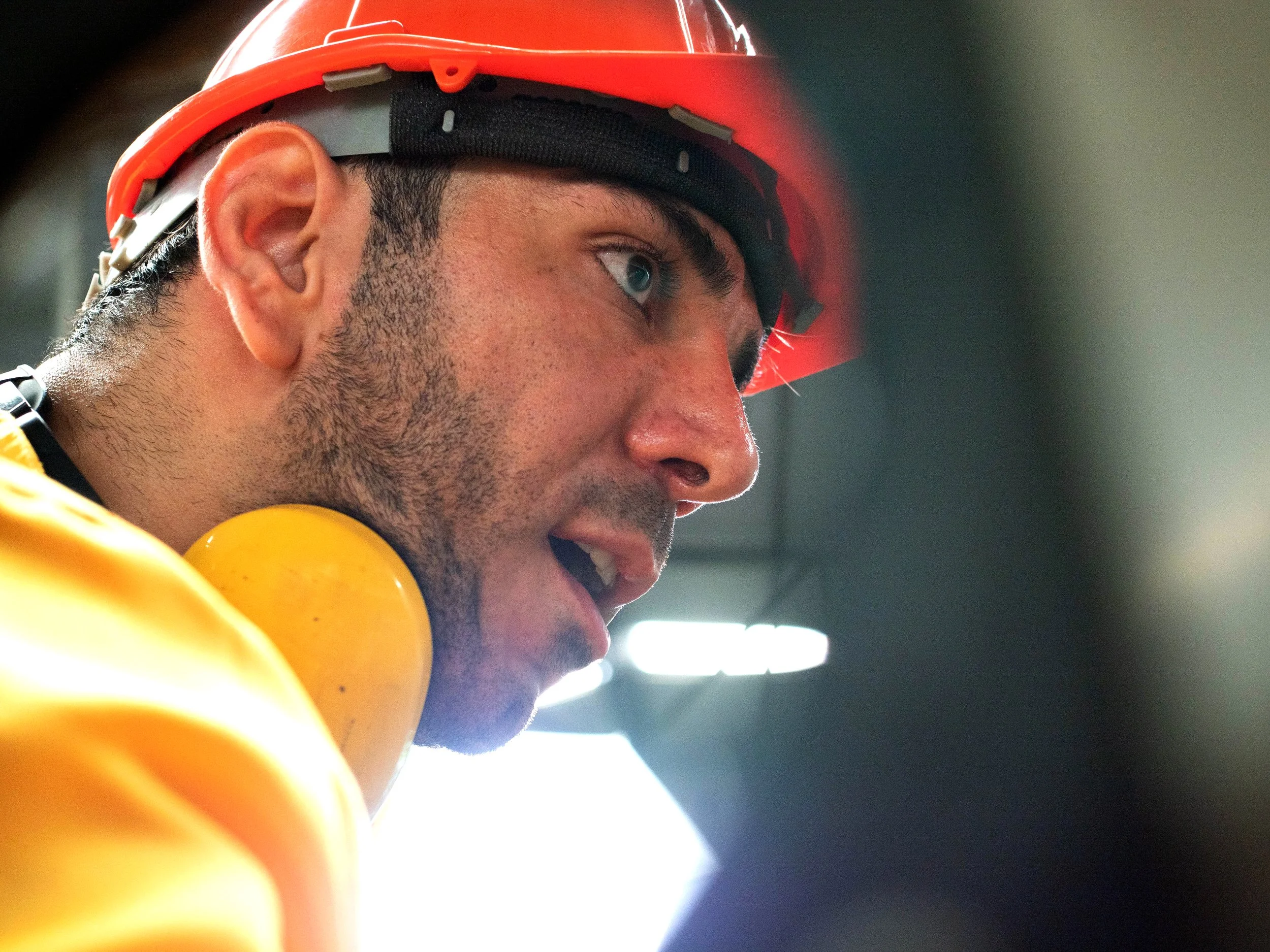 Close-up of a construction worker wearing a red safety helmet and orange high-visibility clothing, with hearing protection around his neck, looking focused.