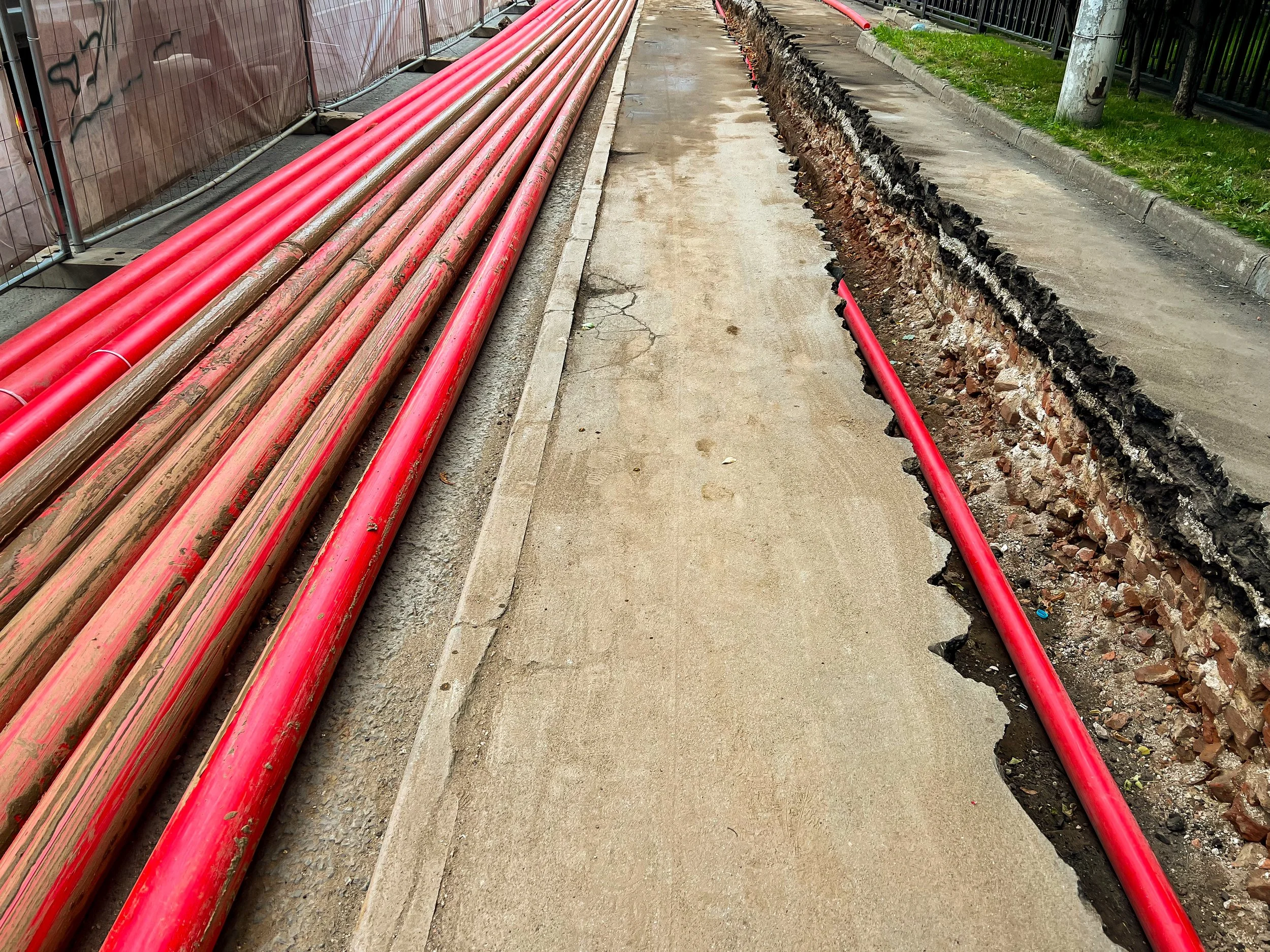 Construction site with red pipes on footpath and an open trench with a single red pipe inside.