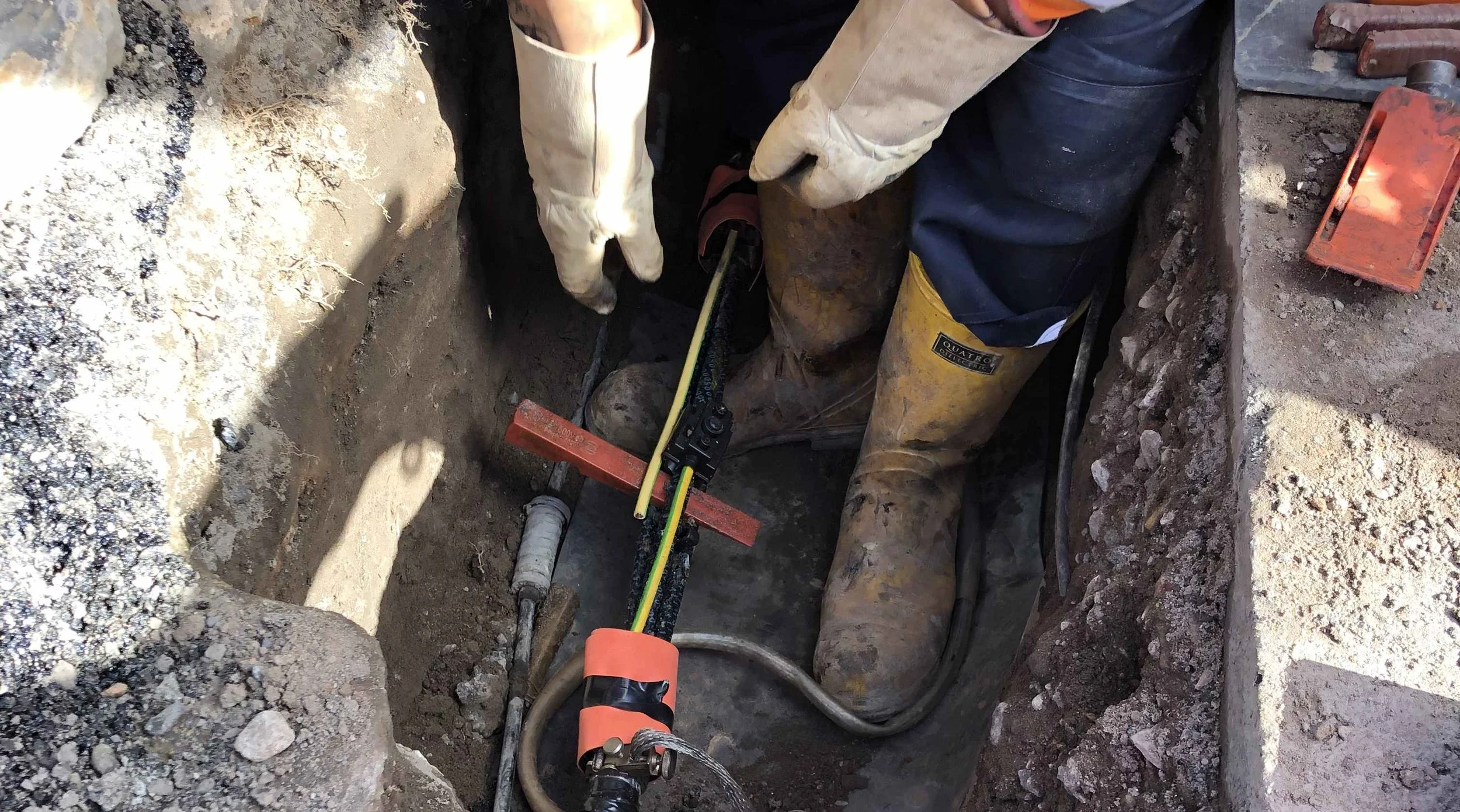 A construction worker wearing yellow rubber boots and gloves is working on electrical cables in a deep hole surrounded by dirt and debris.