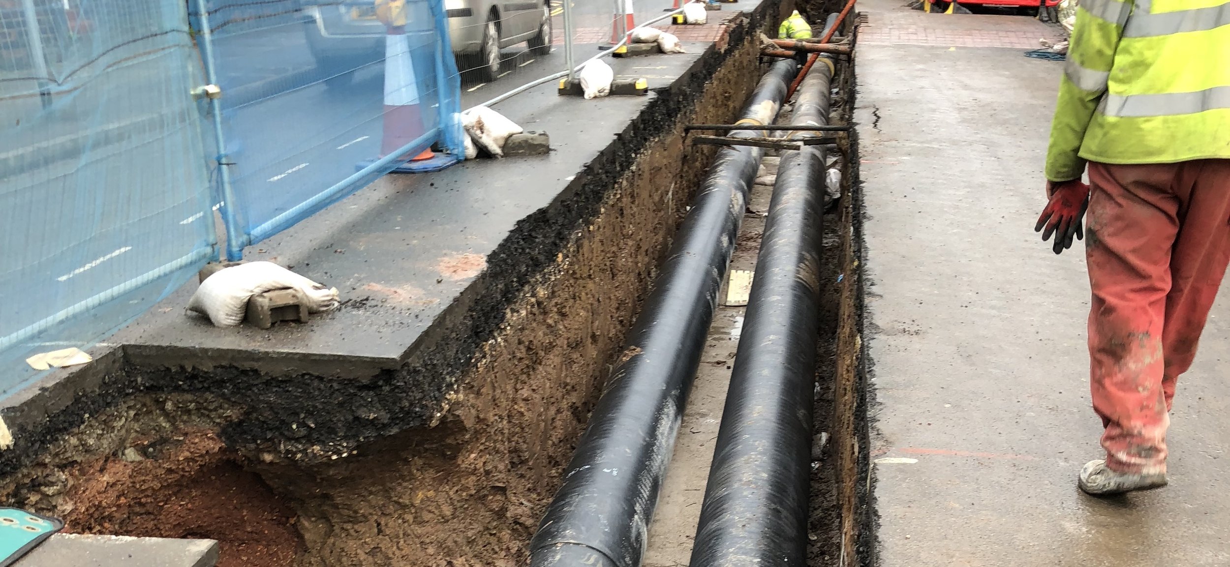 A construction worker standing near an excavation with two large underground pipes on a city footpath.