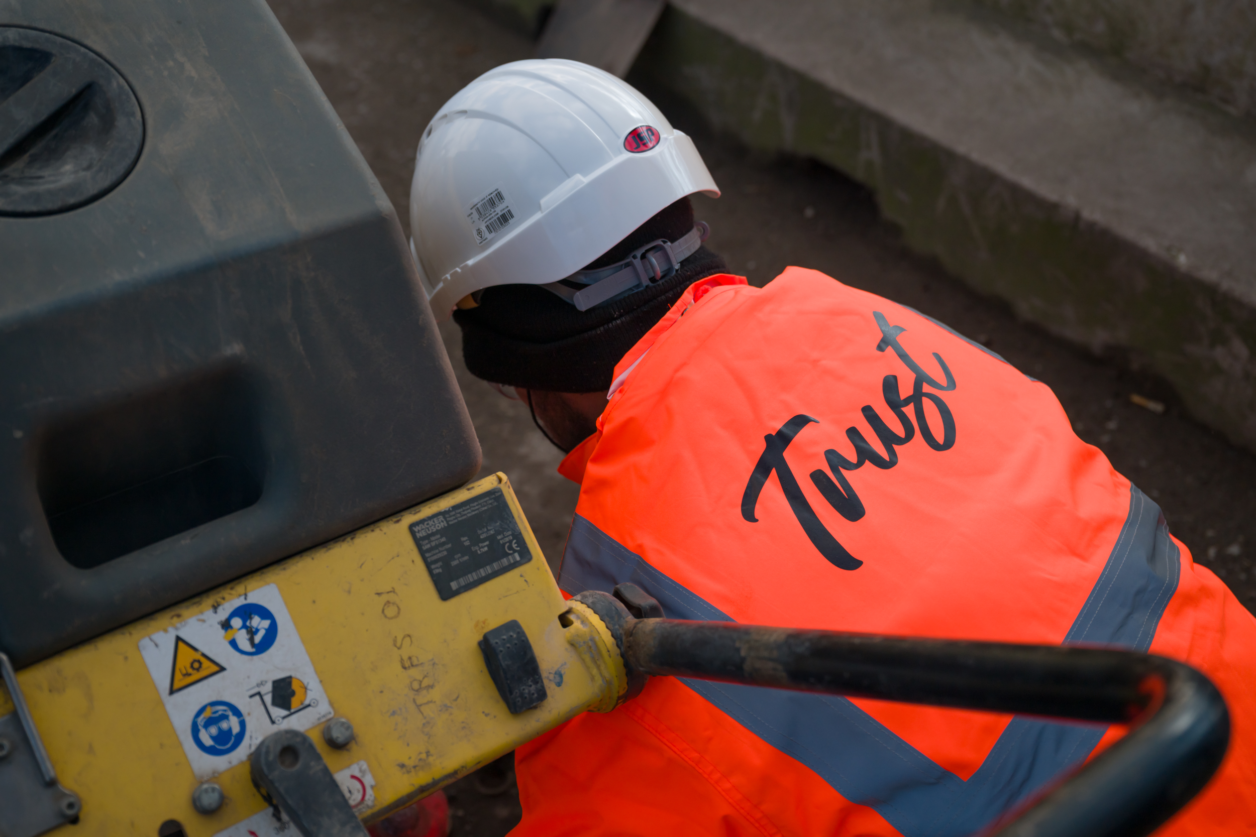 A construction worker in an orange safety jacket and white safety helmet using machinery near concrete steps.