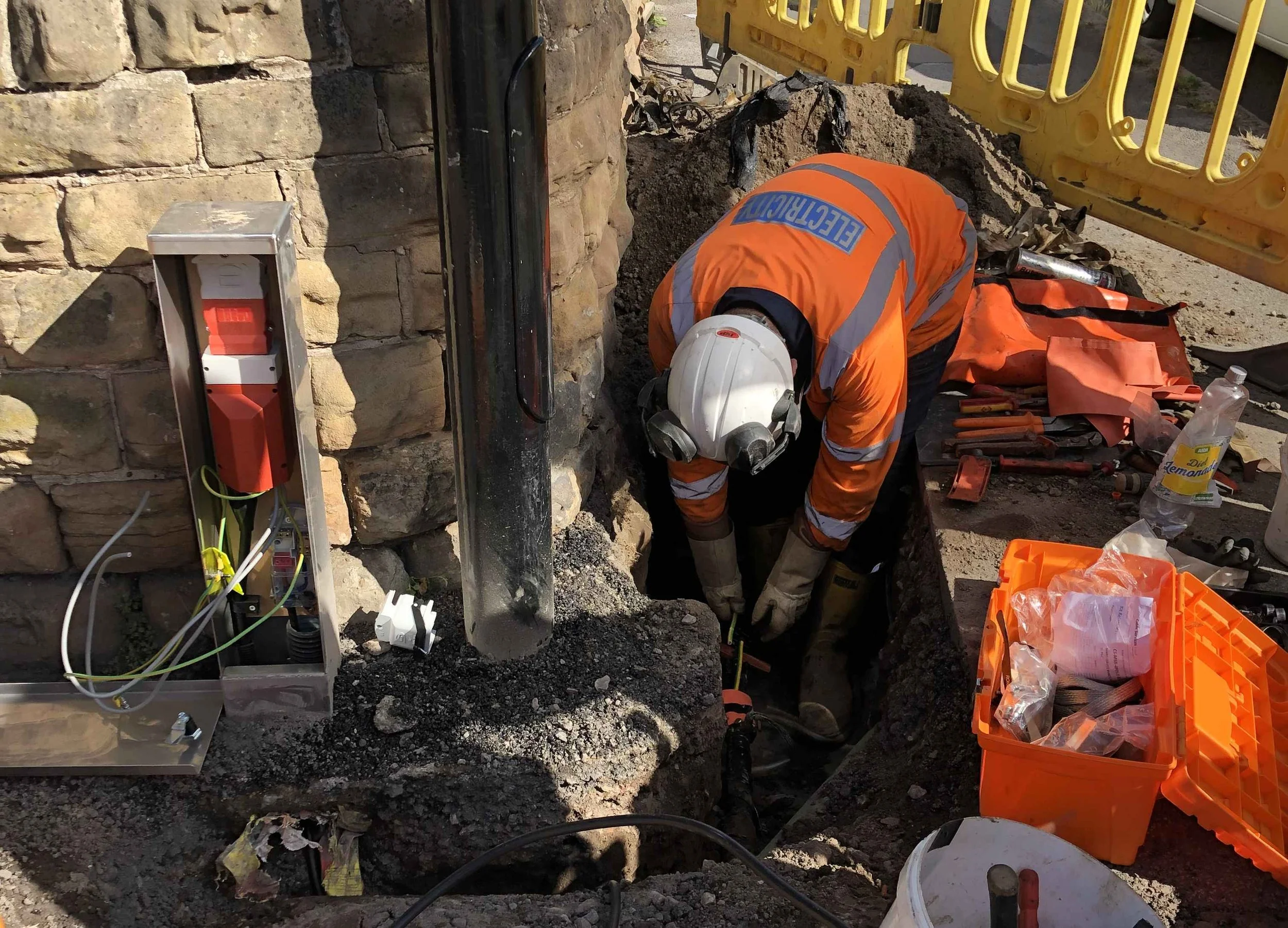 Construction worker in orange safety gear and white helmet working in a trench next to a stone wall with an electrical box and tools nearby.