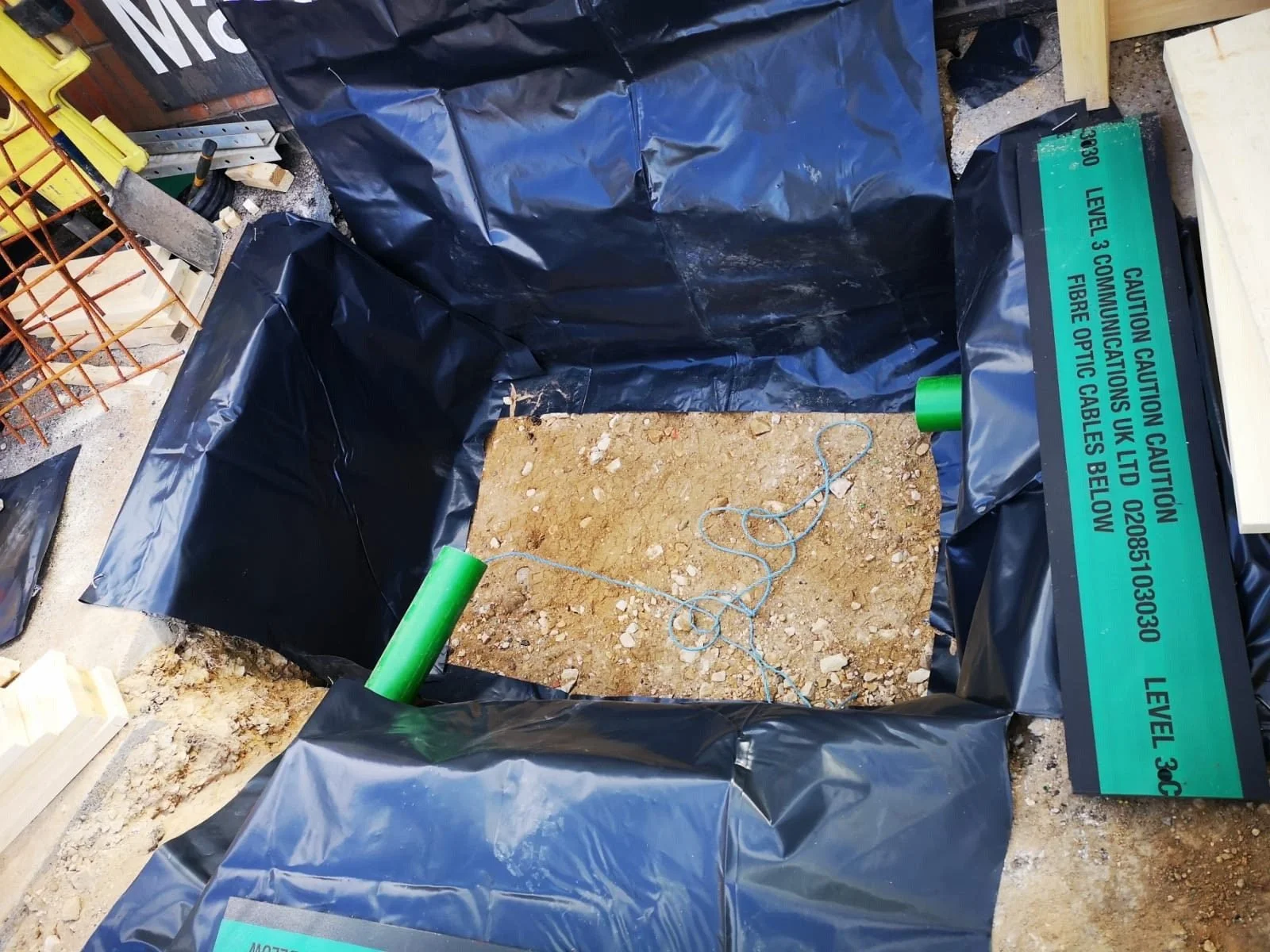 A construction site with a trench or hole lined with black plastic sheeting and two green pipes, one apexed and angled, others extending from the sides. There are warning signs on the covered area indicating caution.