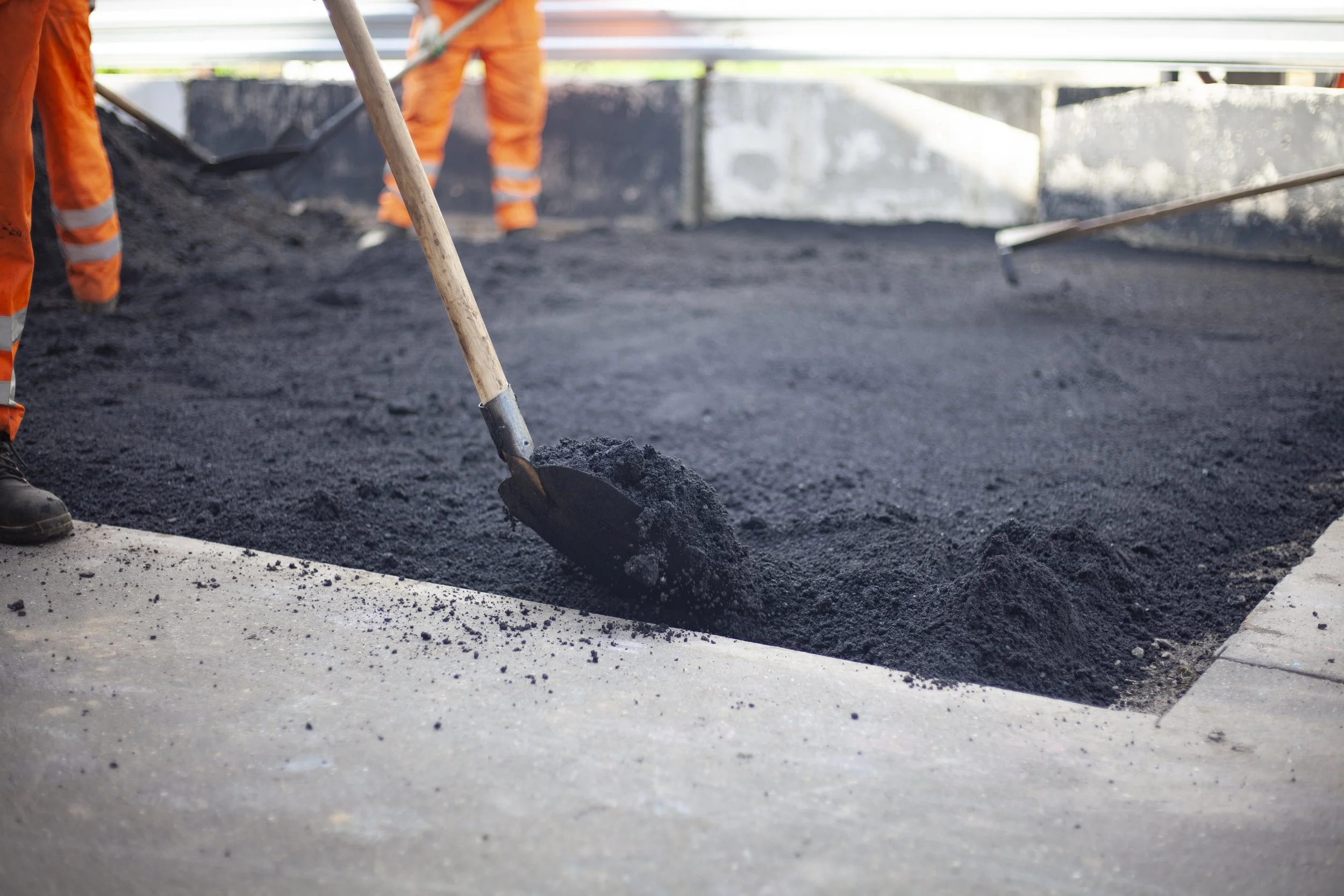 Construction workers paving a road with black asphalt. They are using shovels and wearing orange safety pants.