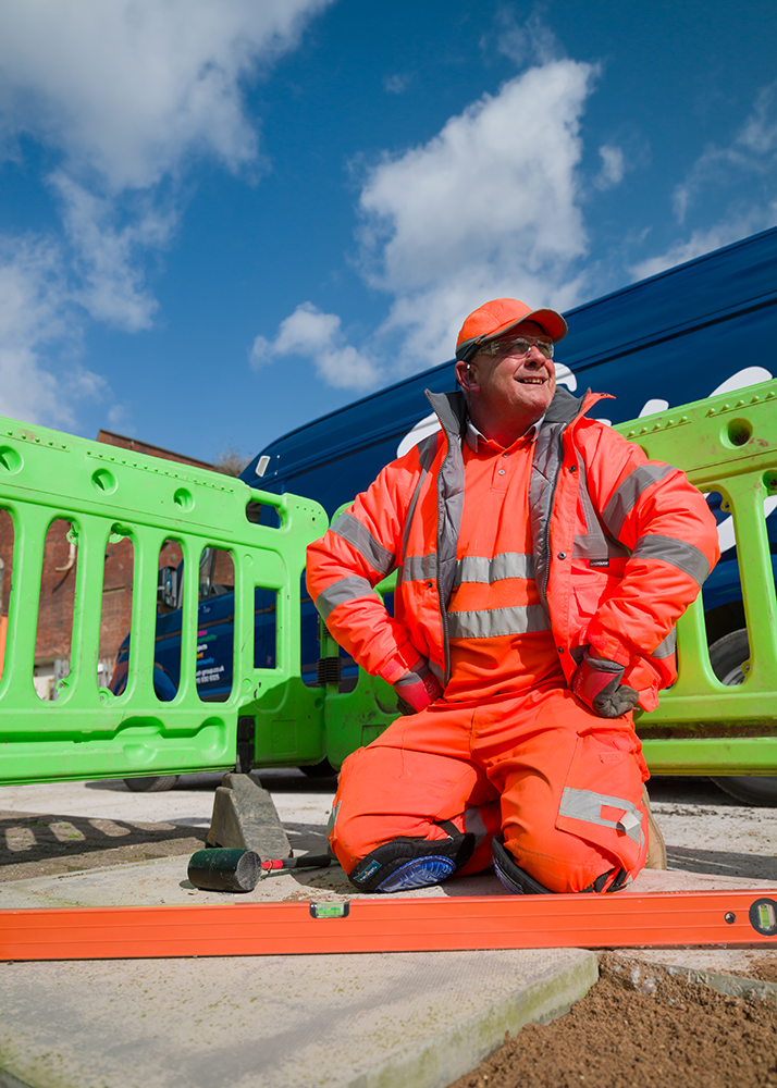 A trust worker laying paving flags behind green barriers against a blue sky.