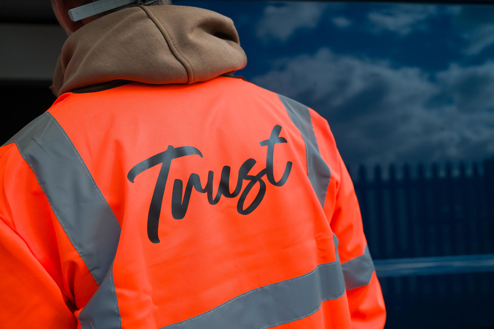 A person wearing an orange high-visibility jacket with gray reflective stripes, with the word 'Trust' written on the back in black script.