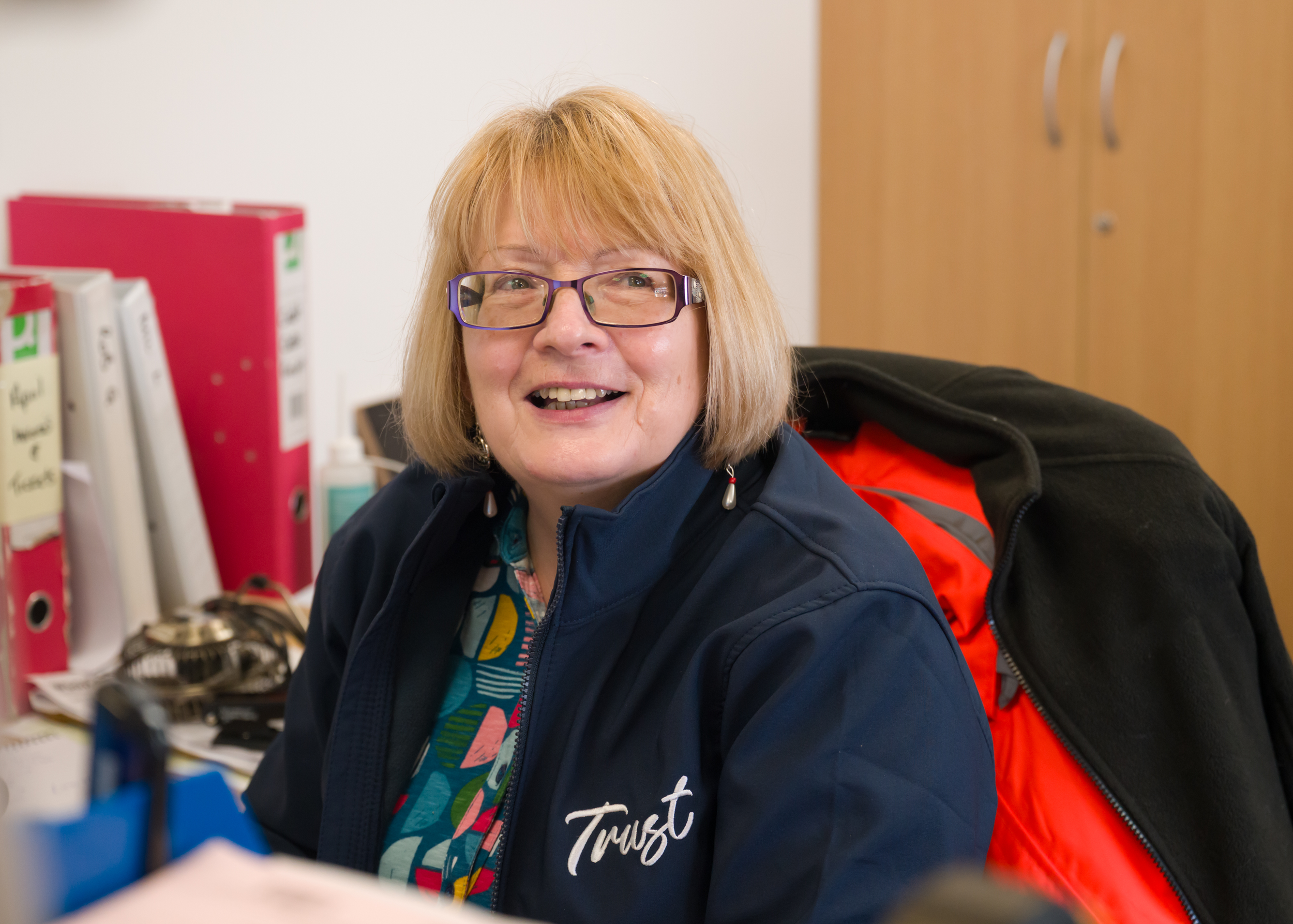Female office staff sitting at a desk wearing a Trust branded blue jacket