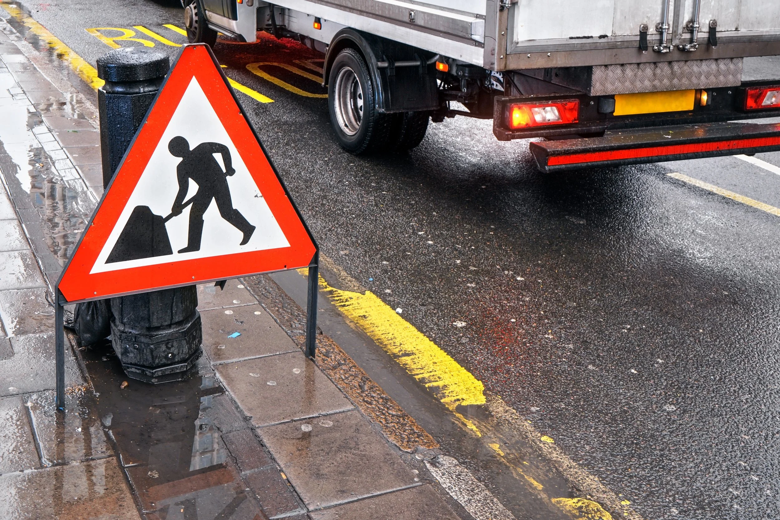 Wet street with a construction worker road work sign on the sidewalk near a parked truck.