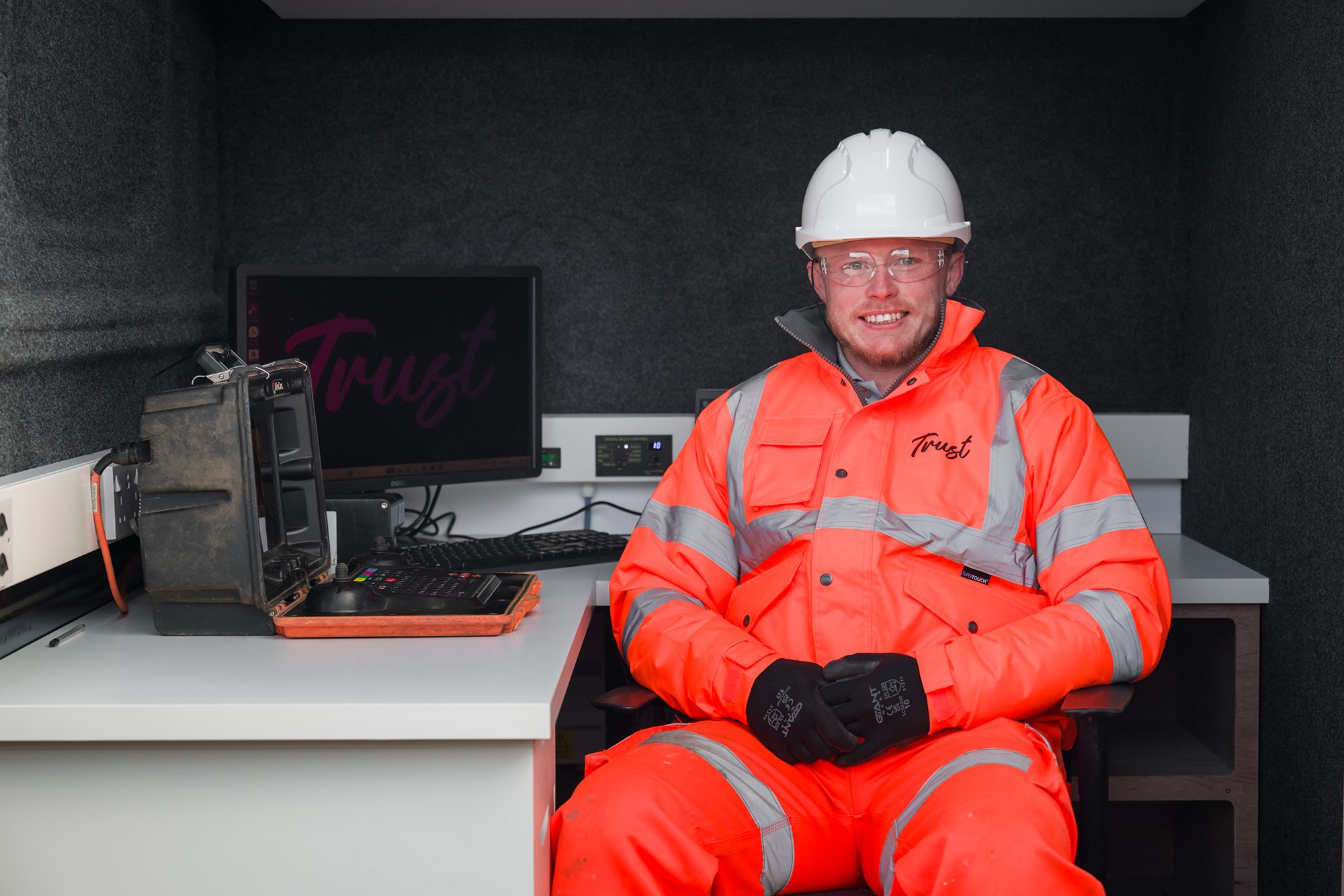 Male Trust worker sat at a desk on a drainage division van next to a computer wearing a white hard hat, safety glasses and orange hi-vis clothing smiling at the camera.