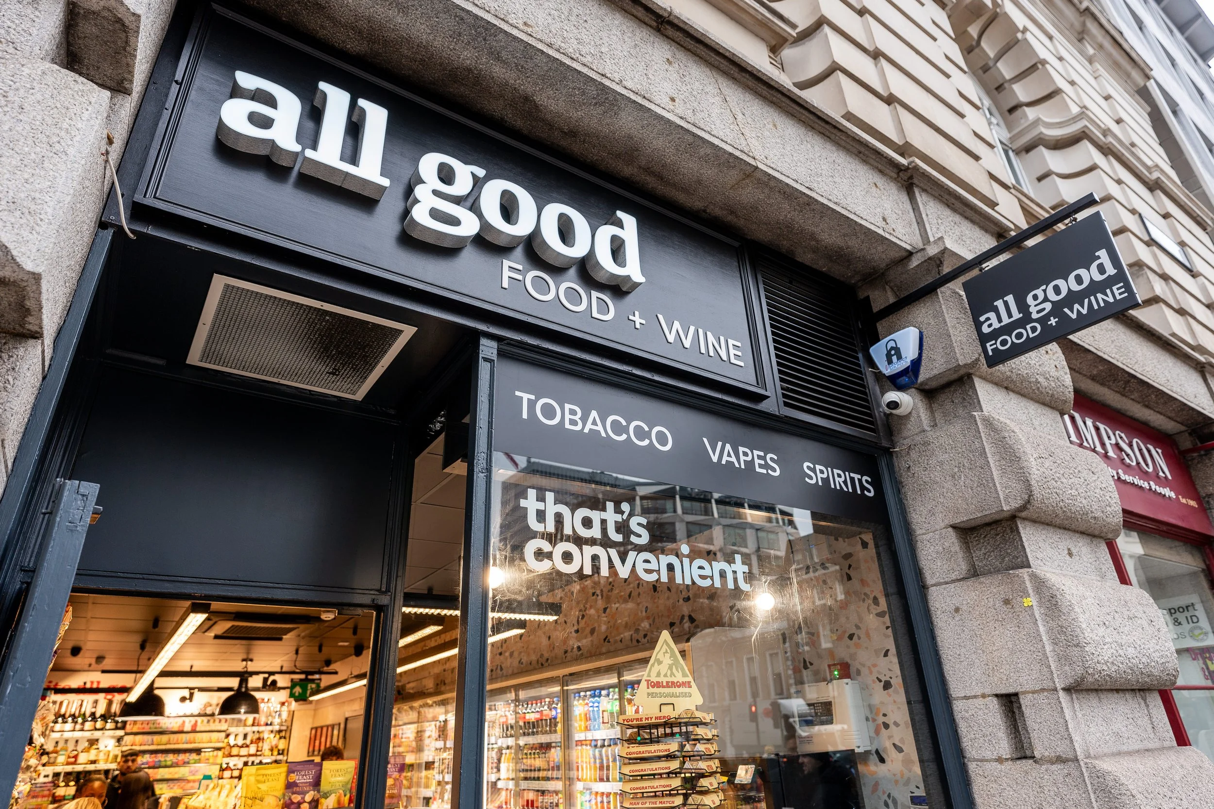 Exterior of a store named all good offering food and wine, with signs indicating tobacco, vapes, and spirits inside.