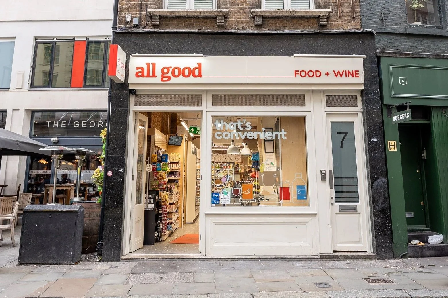 Front view of a small convenience store named 'all good' with a sign that reads 'FOOD + WINE' and a window displaying bottled beverages and grocery items inside.
