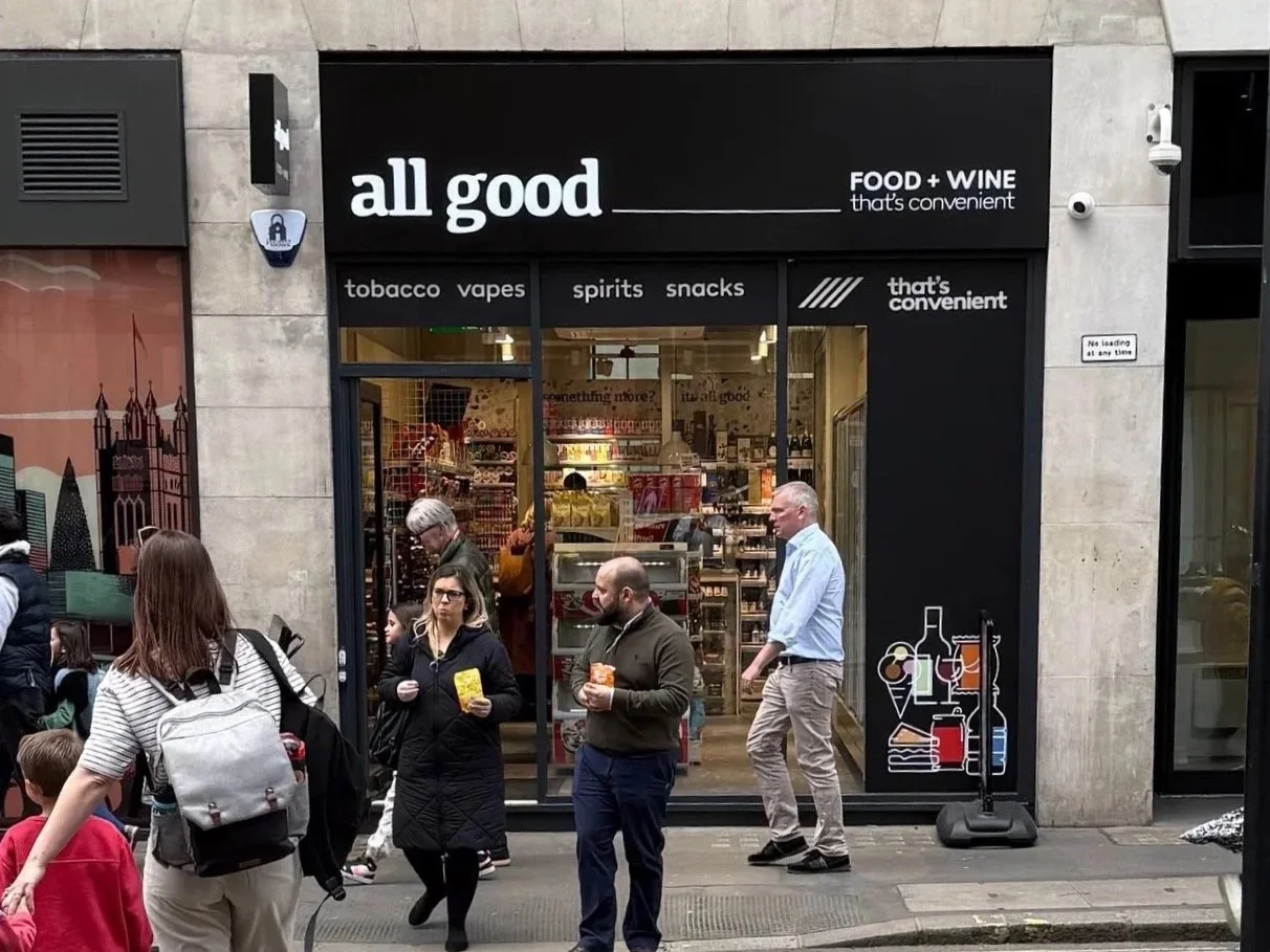 Street scene outside a shop named 'all good' that sells tobacco, vapes, spirits, snacks, and wine. Pedestrians are walking by, some holding snacks. The shop has large glass windows displaying products inside. Signage advertises convenience and a no loading zone at the curb.