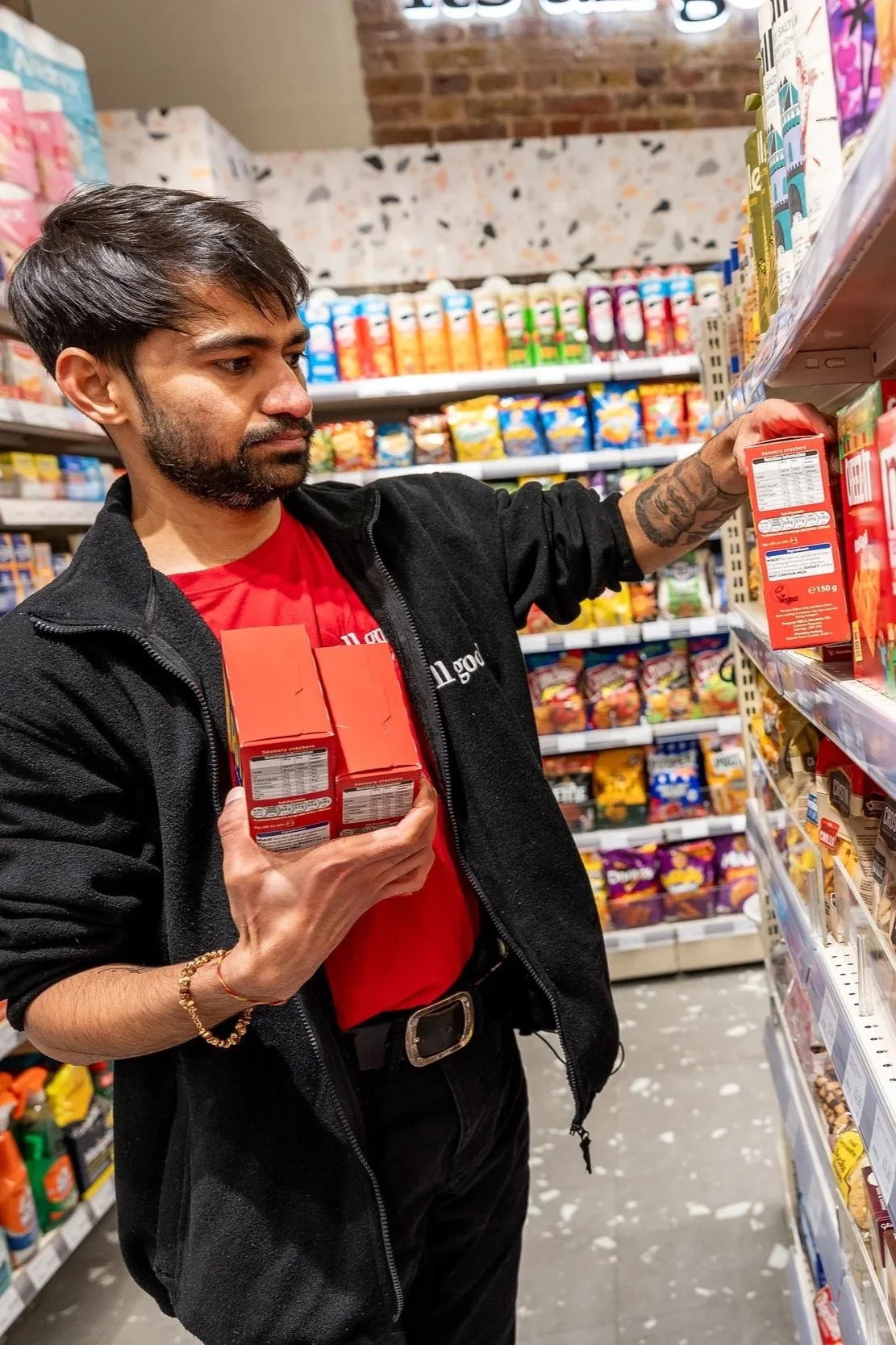 A man shopping in a grocery store, holding three boxes of a product, reaching for another on the shelf.