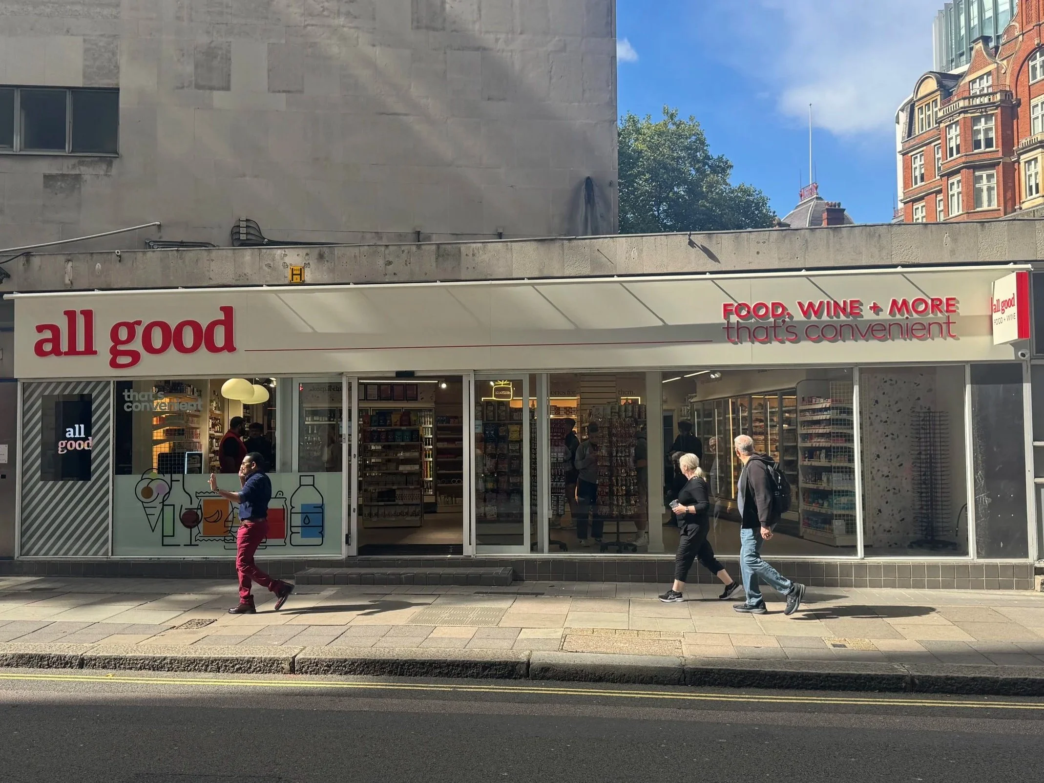 A storefront named 'all good' with signage that says 'food, wine + more, that's convenient.' Glass doors show shelves of products inside and three people walking on the sidewalk in front.