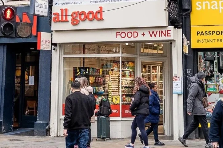 People walking past a small store named All Good that sells food and wine, with various snacks visible inside, on a busy street.