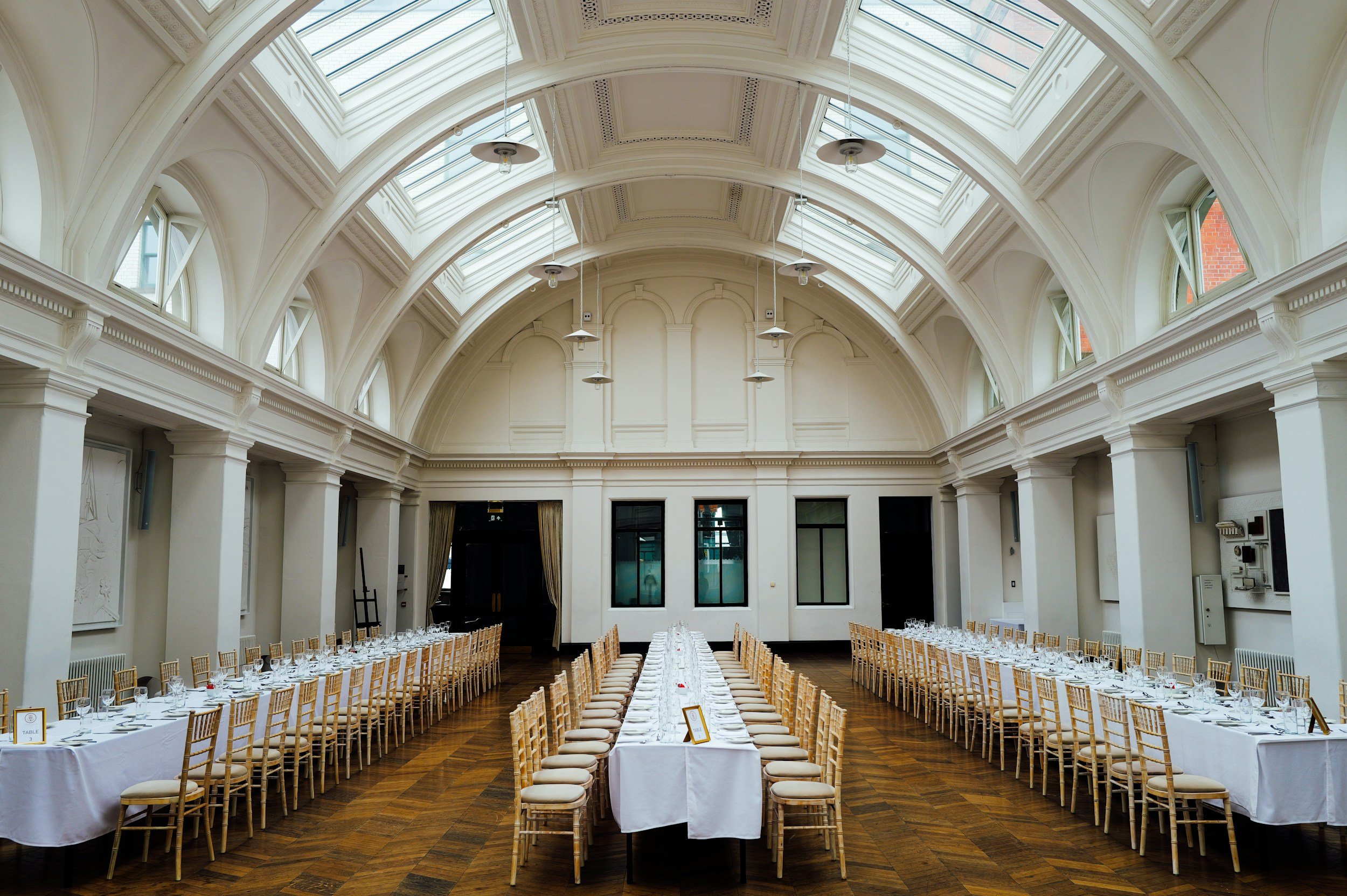 Elegant banquet hall with high vaulted ceiling, large skylights, and long tables set with white tablecloths, glassware, and gold chairs, ready for a formal event.