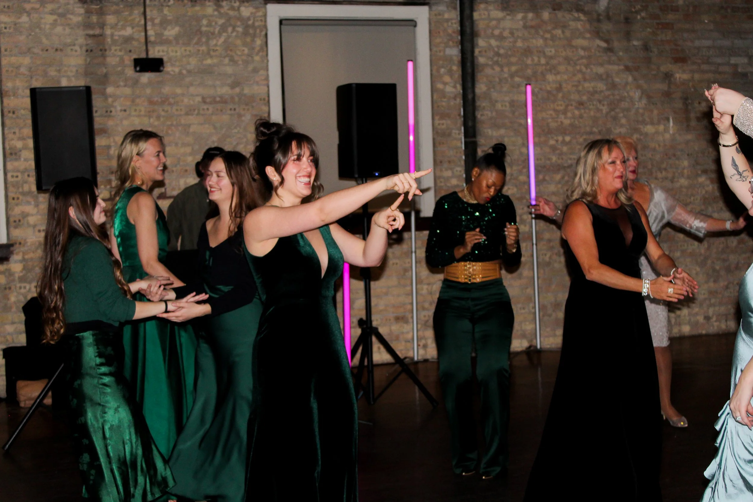 Women dancing and celebrating at an indoor party or event with brick wall background, some holding light glow sticks.