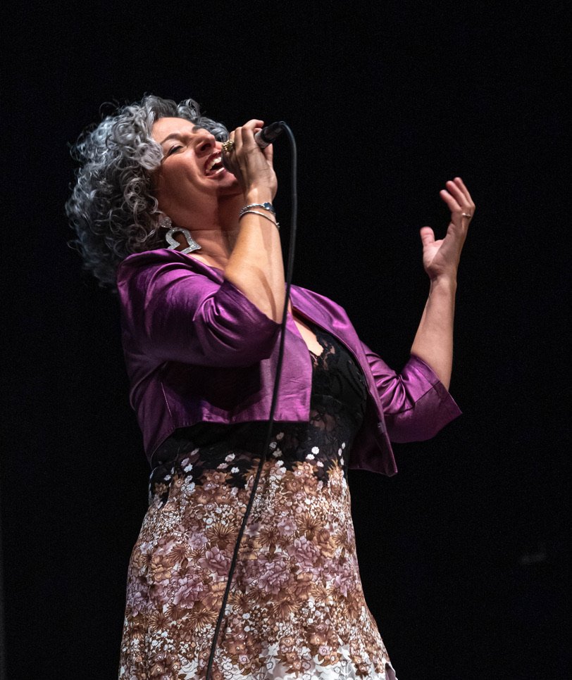 Anna Chiara Farneti che canta con microfono, capelli ricci grigi, indossa giacca viola e gonna con motivo floreale, sfondo nero.