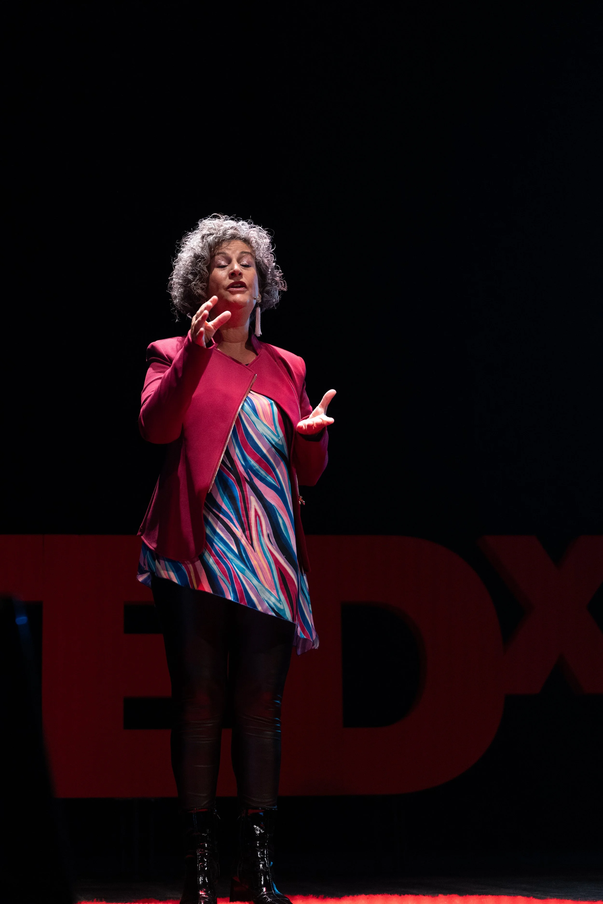 Anna Chiara Farneti  indossa una giacca rossa e un vestito con motivo multicolore, che sta parlando o presentando su un palco di fronte a un pubblico.