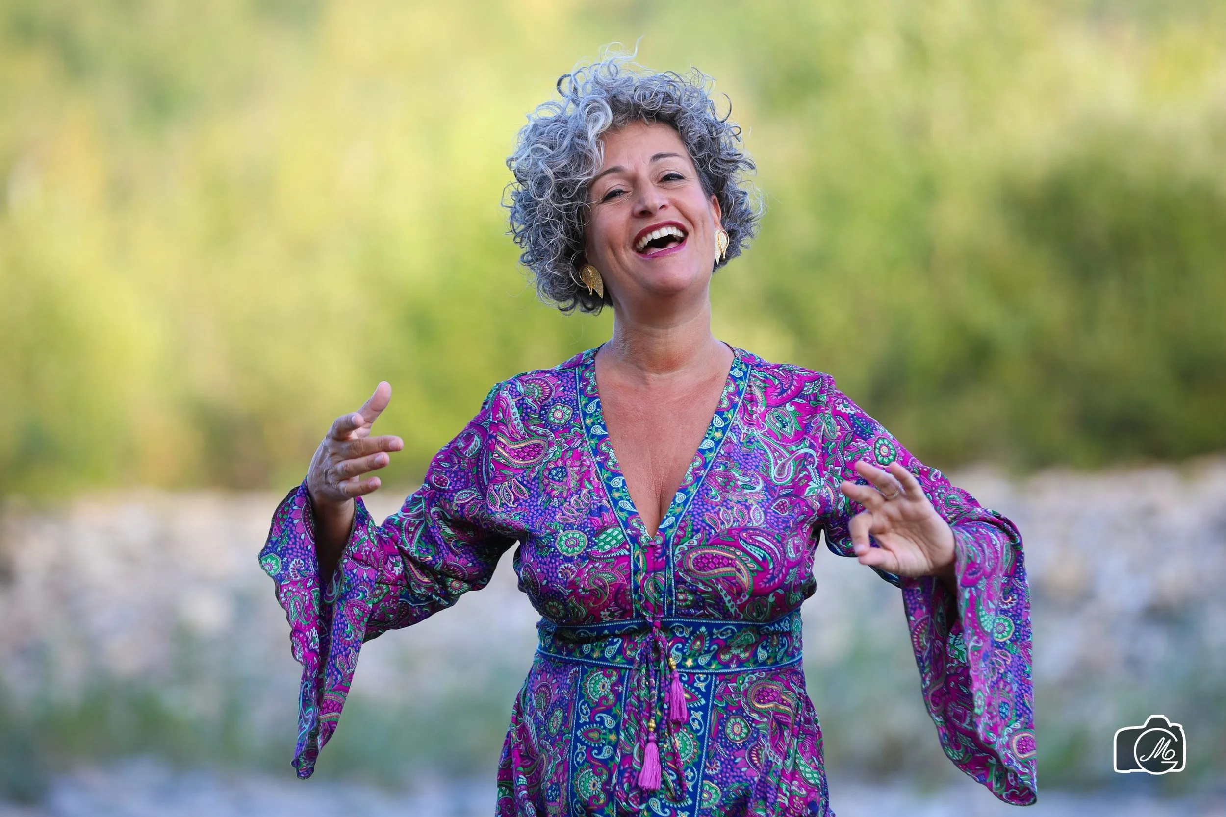Una donna con capelli ricci grigi che sorride mentre si trova all'aperto con sfondo di natura verde.