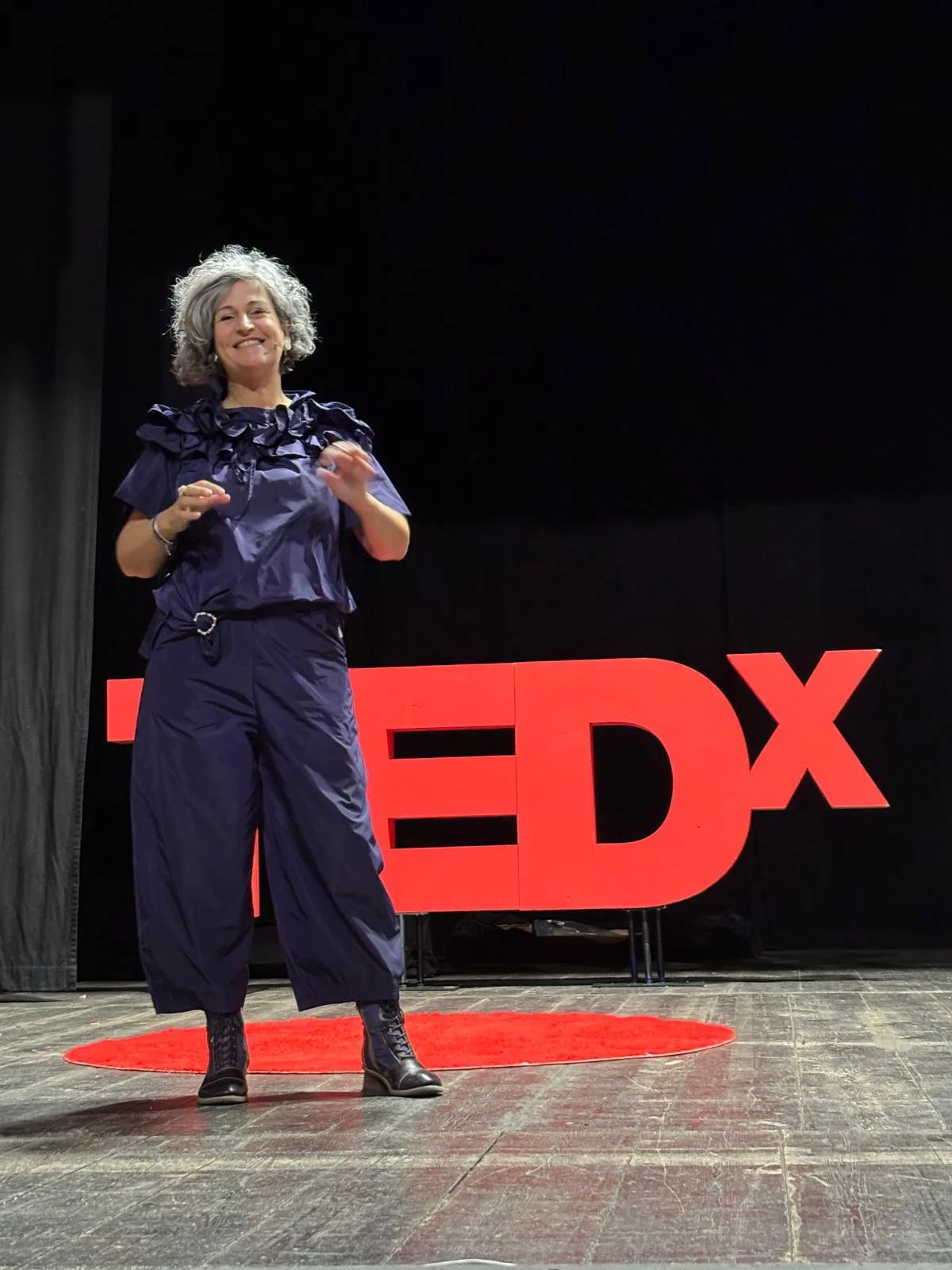 Anna Chiara Farneti che parla sul palco di un evento TED, con il logo TEDX rosso sullo sfondo e un tappeto rotondo sotto i piedi.