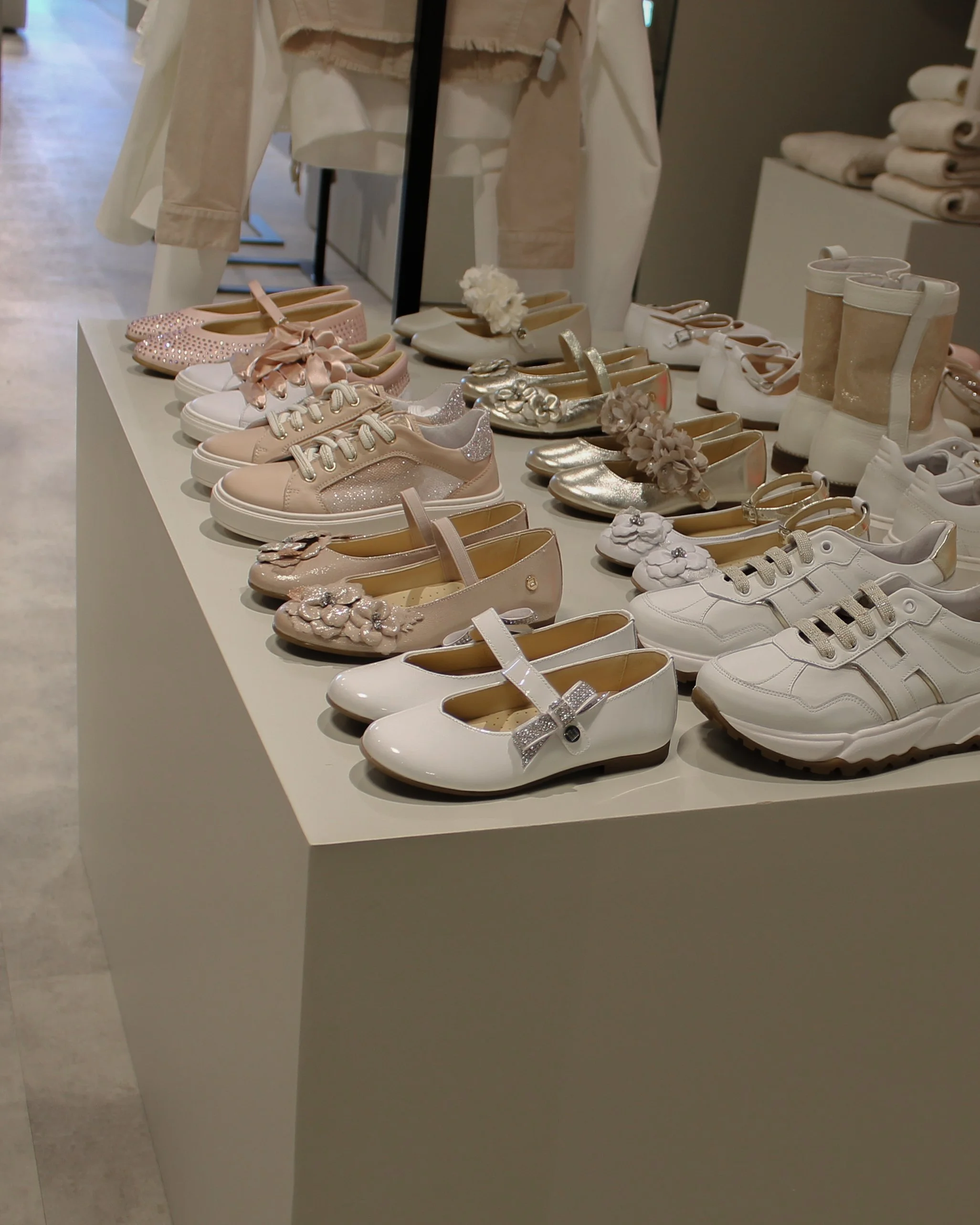 Een uitstalling van kinderschoenen, waaronder sneakers, ballerina's en laarzen in roze, wit en metallic kleuren, op een witte kubus in een kledingwinkel.