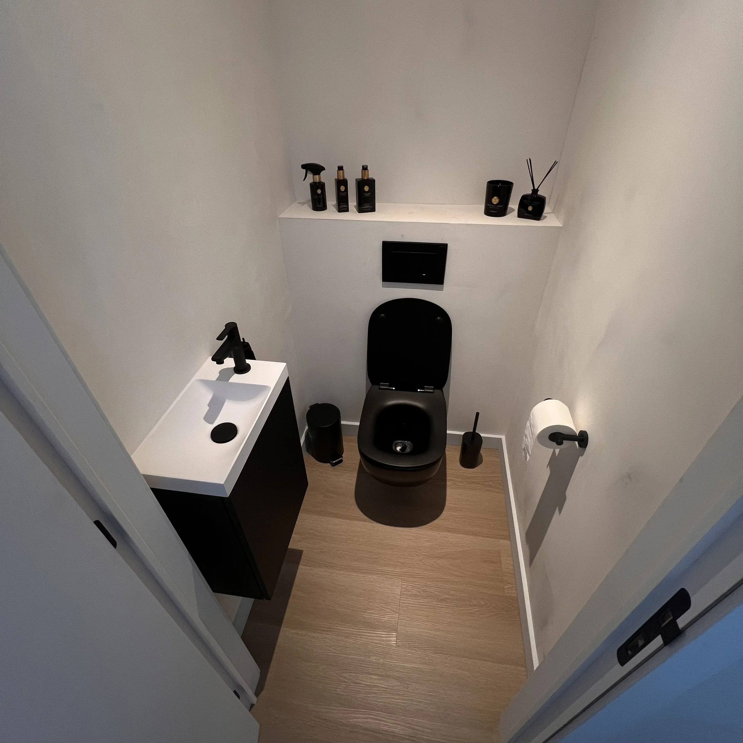 Minimalistische zwart-witte badkamer met een zwart toilet, een kleine zwarte wastafel met zwarte kraan, toiletpapier aan de muur en zwarte decoratieve voorwerpen op een plank boven het toilet.