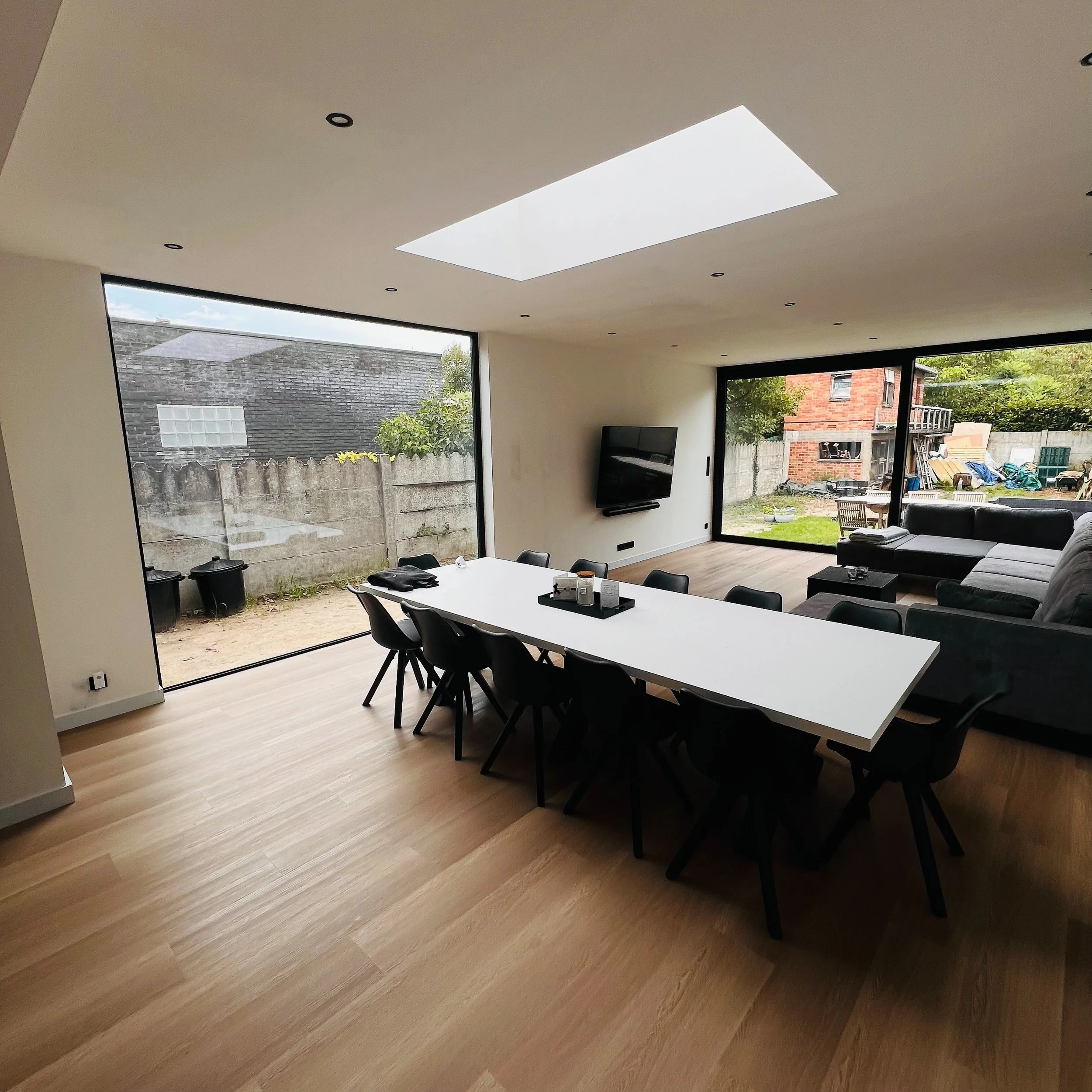 Moderne eet- en woonkamer met grote ramen, houten vloeren, een witte eettafel met zwarte stoelen, een aan de muur gemonteerde tv en een gezellige zithoek met een grijze hoekbank, met uitzicht op de achtertuin.
