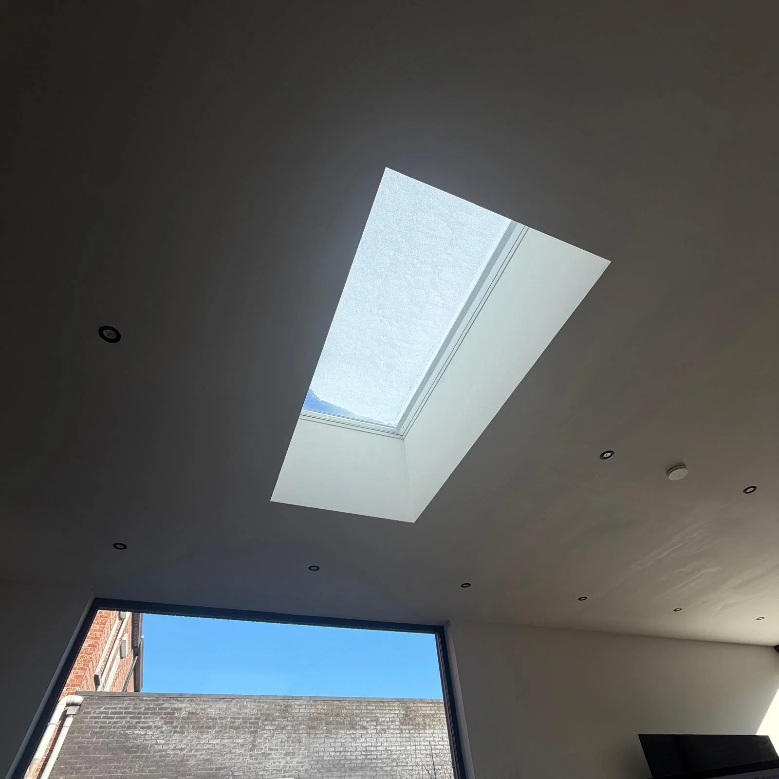 Binnenopname van een kamerplafond met een groot rechthoekig dakraam dat natuurlijk licht binnenlaat, en een raam met uitzicht op een bakstenen gebouw buiten.