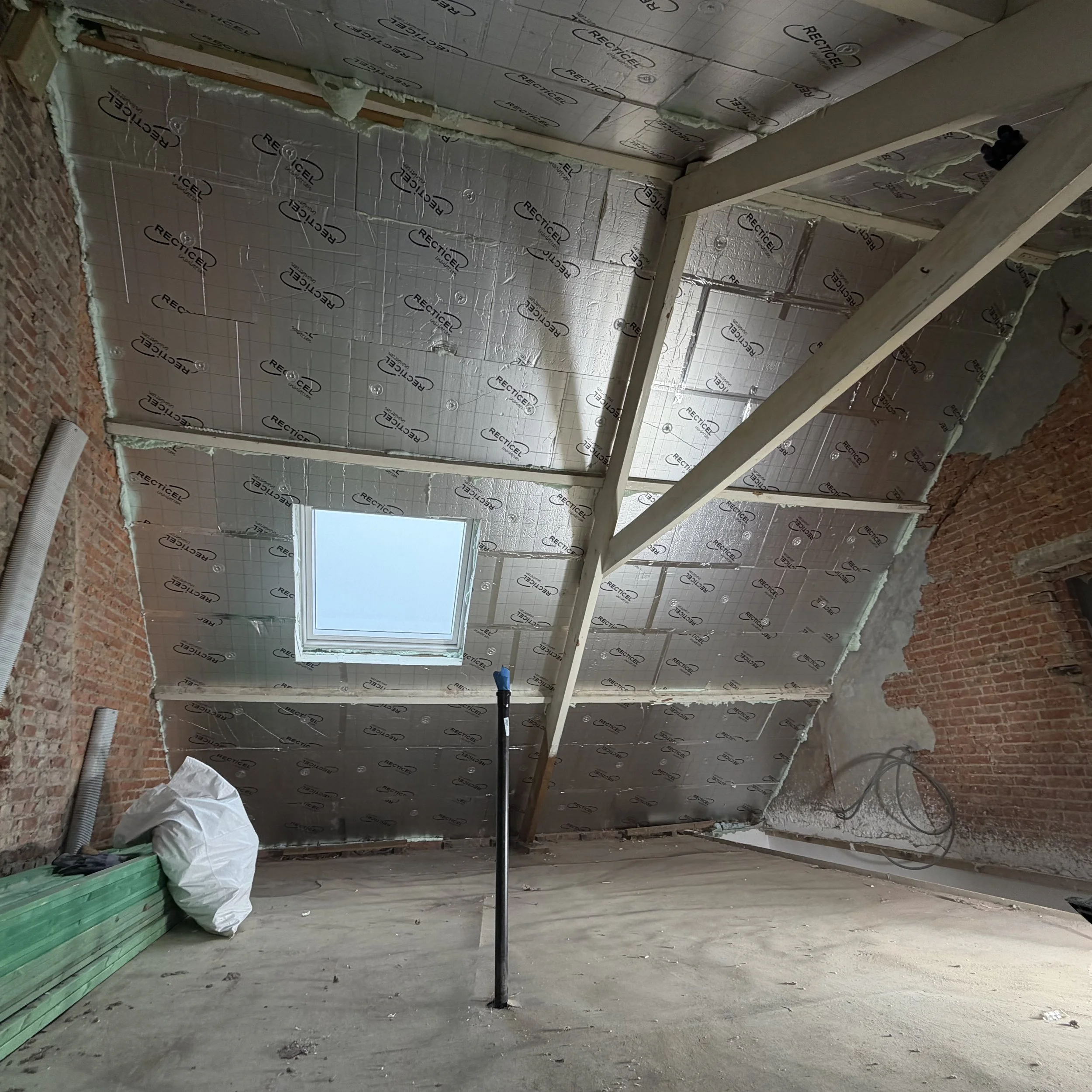 Een onafgewerkte zolderkamer in aanbouw, met isolatie aangebracht op het schuine plafond en een dakraam. Bouwmaterialen en gereedschap zijn zichtbaar en de ruimte is omgeven door bakstenen muren.