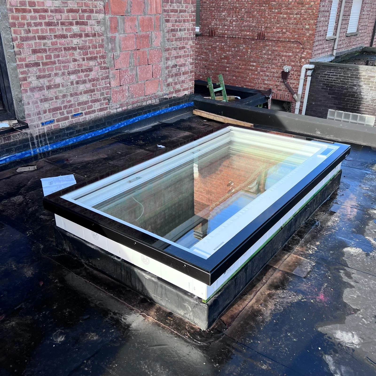 Een rechthoekig dakraam, geplaatst op een plat dak, omgeven door zwart dakbedekkingsmateriaal met aangrenzende bakstenen muren.