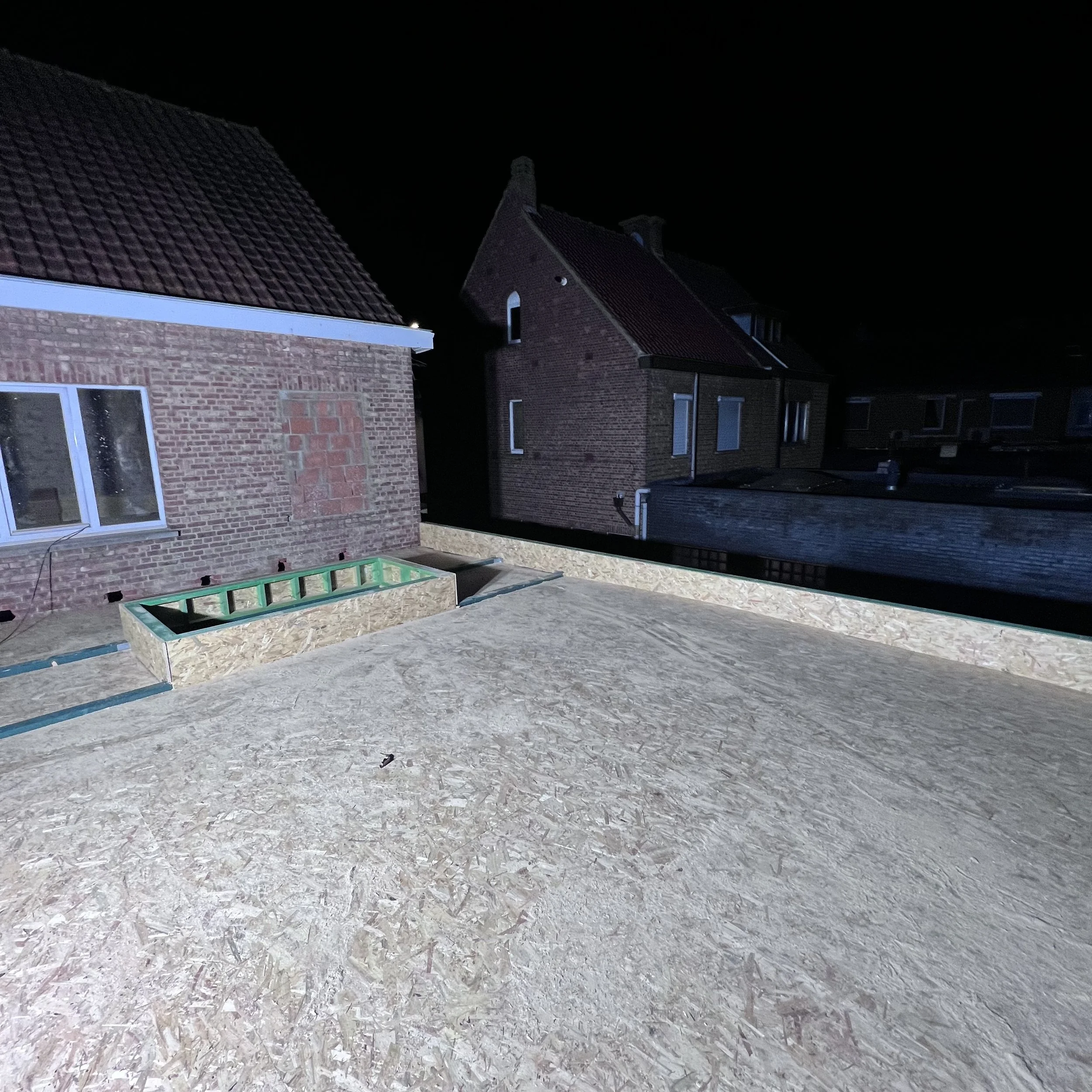 Een dak in aanbouw met multiplex beplating, naburige bakstenen huizen en een raam met glasplaten.