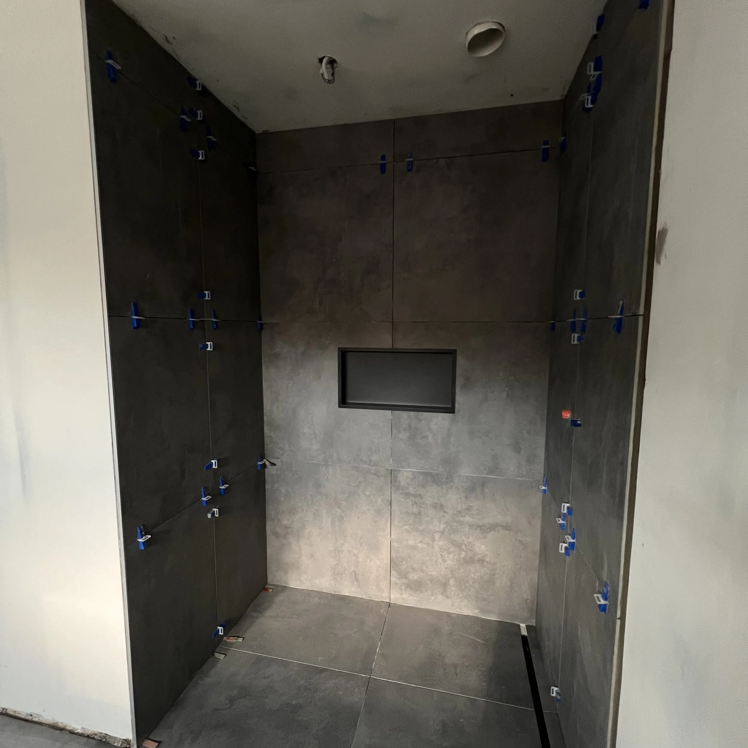 Een douche met donkergrijze tegels, een klein rechthoekig raam en nog aanwezige blauwe tegelafstandhouders.