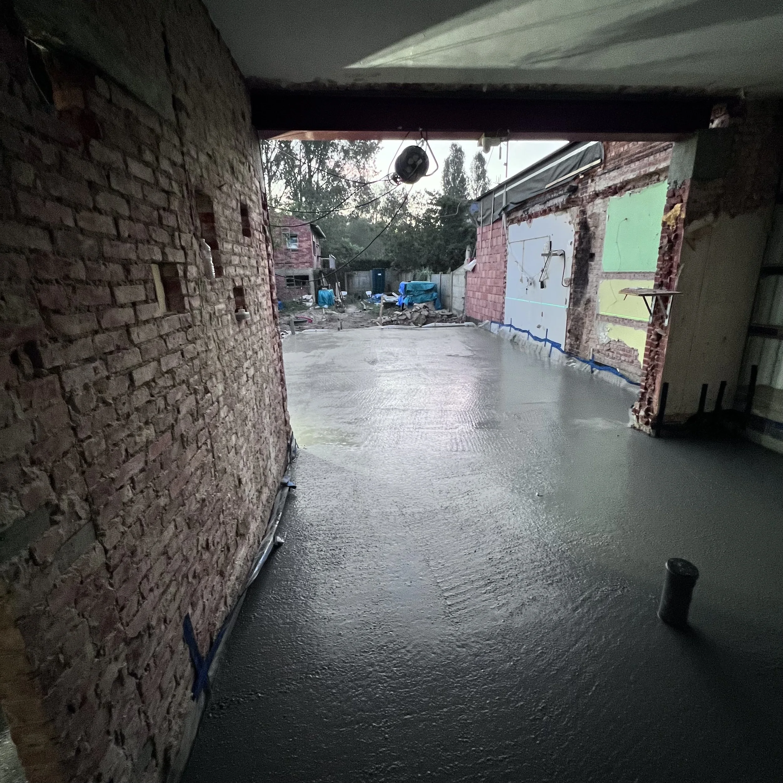 Een pas gestorte betonnen vloer op een bouwterrein, omgeven door bakstenen muren en gedeeltelijk overdekt door een dak. Op de achtergrond zijn bouwmaterialen en hekwerk te zien, met daarachter bomen.