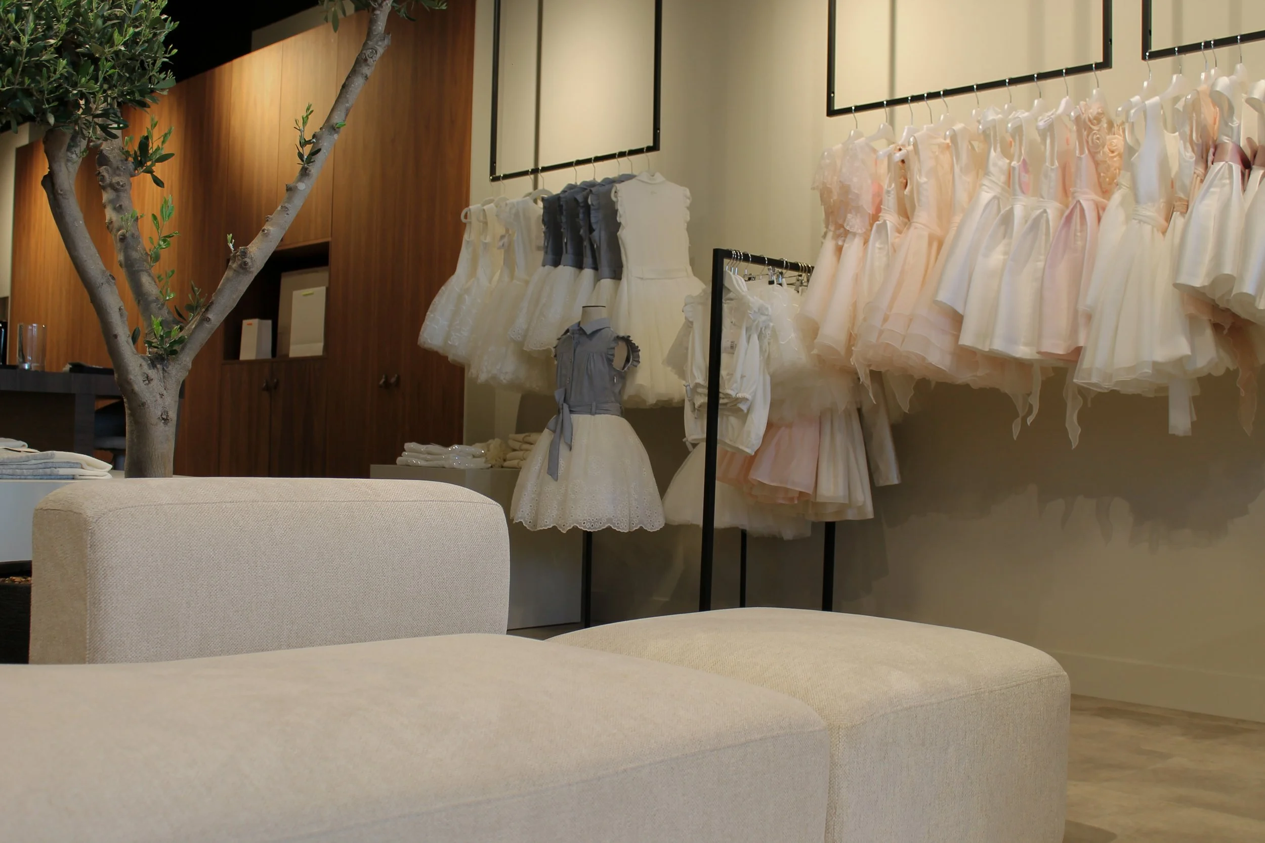 Kinderjurkjes en -outfits tentoongesteld in een winkel, in crème-, wit- en roze tinten, met een beige bank op de voorgrond.