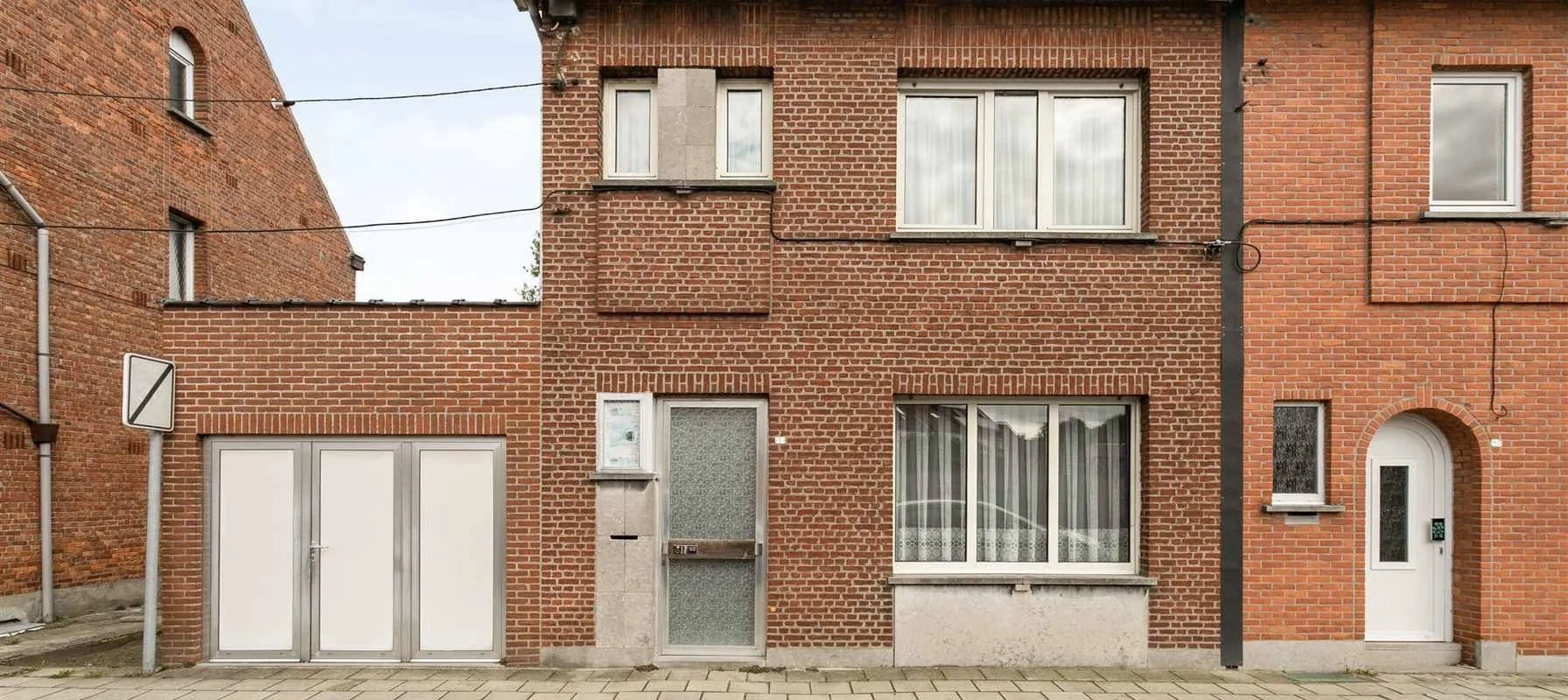 Voor renovatie - Een bakstenen woonhuis met diverse rechthoekige ramen, een witte deur met een gebogen kozijn, een smalle vensterbank en een aangebouwde garagedeur. Bovengronds lopen elektriciteitskabels.