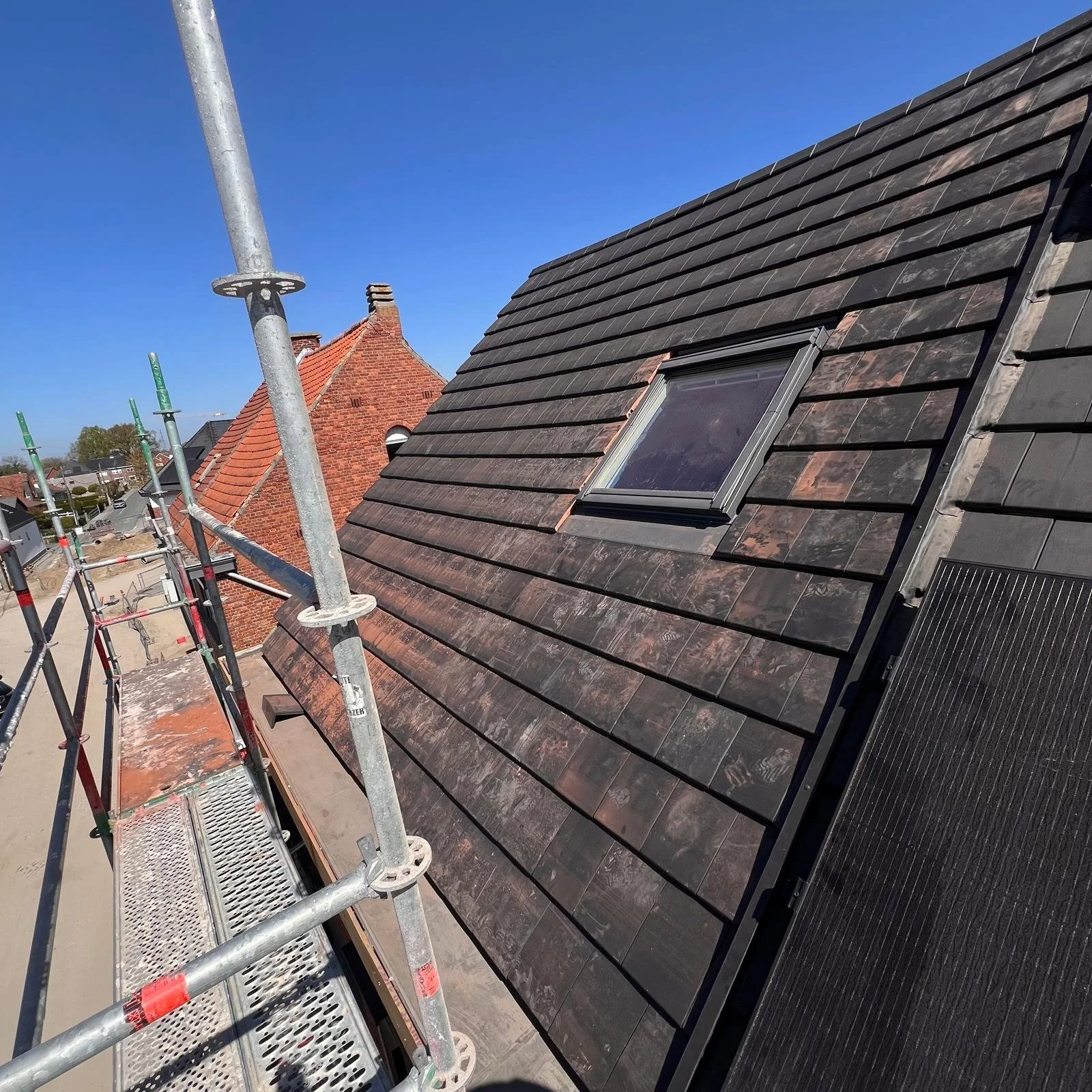 Een dak met donkere dakpannen en een dakraam; aan de linkerkant staat een steiger en tegen een helderblauwe lucht zijn de omliggende bakstenen daken zichtbaar.