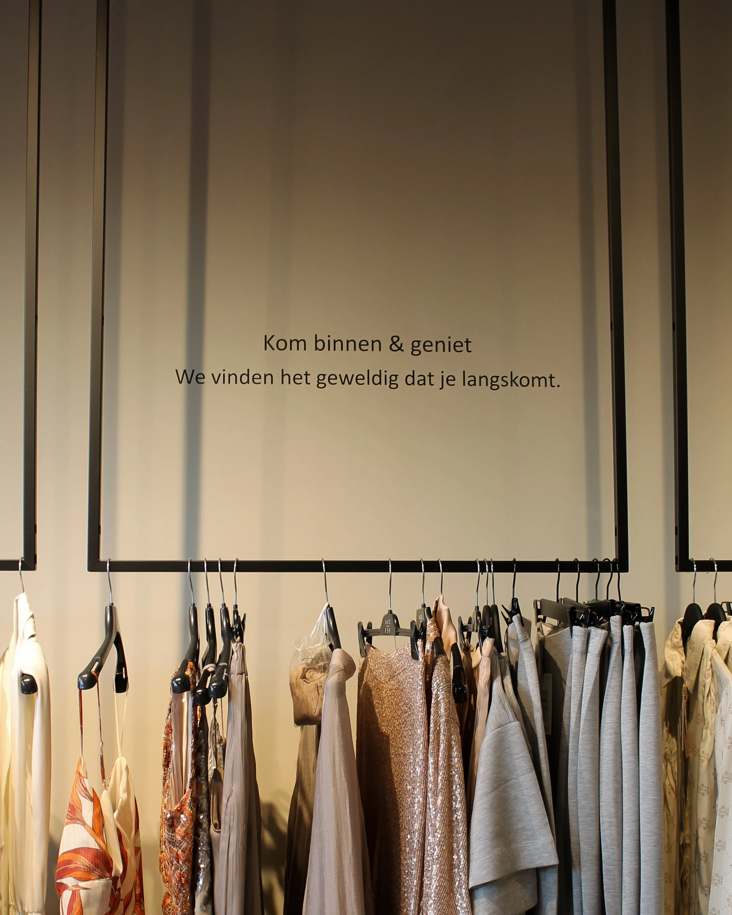 Een kledingrek met de tekst: 'Kom binnen en geniet. We vinden het geweldig dat je er bent.'