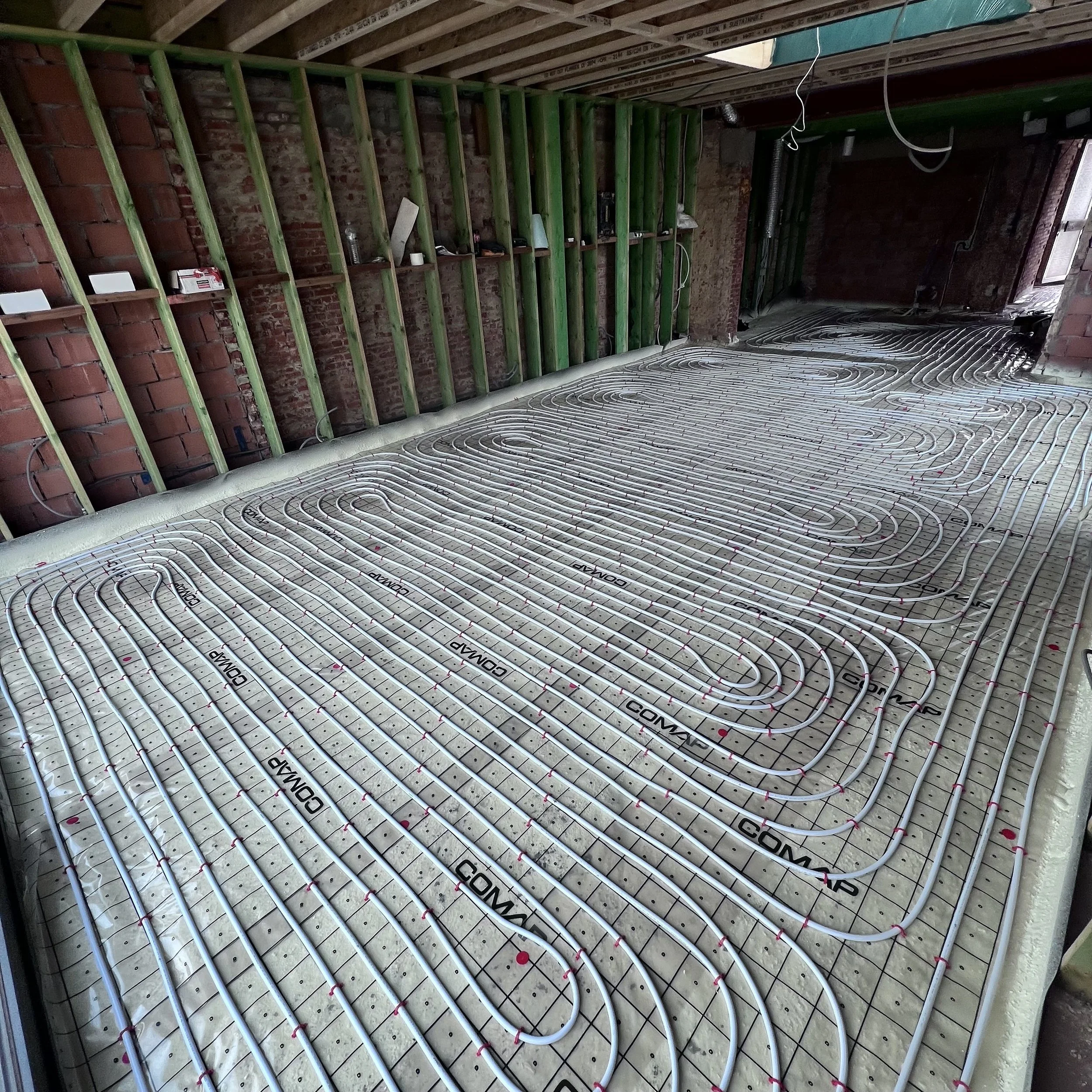 Een ruimte in aanbouw met vloerverwarmingsbuizen in de ondervloer, groene houten wandconstructie, zichtbare bakstenen muren en een onafgewerkt plafond.