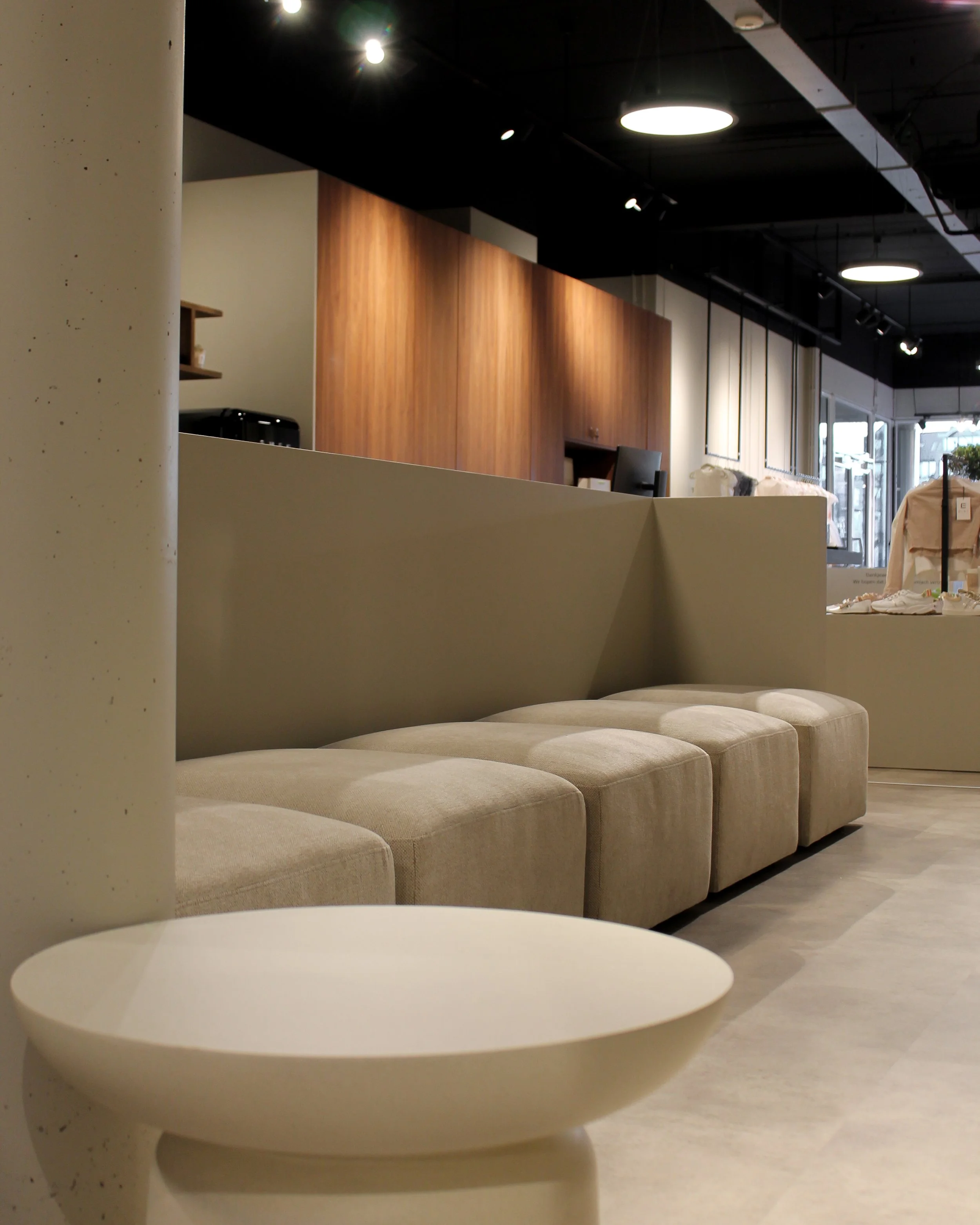 Een modern, minimalistisch interieur met beige gestoffeerde banken, een kleine ronde witte tafel op de voorgrond en warme houten kast achter de toonbank.