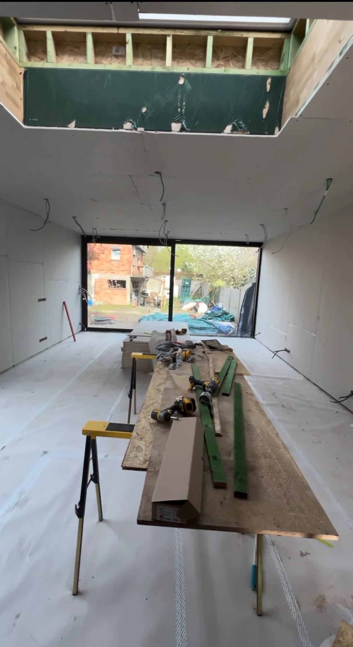 Een binnenruimte in aanbouw, met gedeeltelijk geplaatste gipsplaten, hangende draden, gereedschap op een werkbank en een groot raam dat uitzicht biedt op de buitenomgeving.