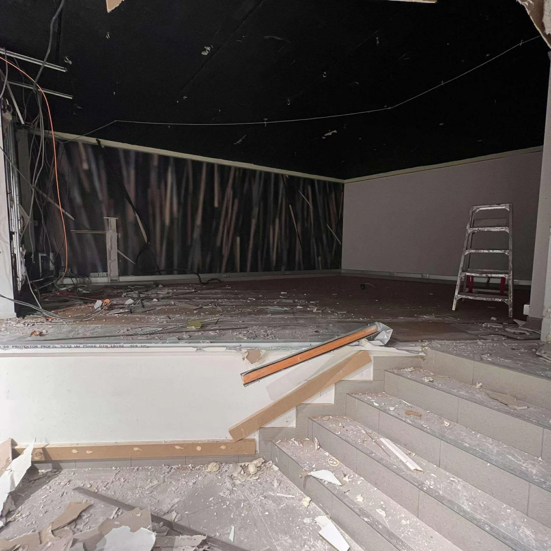 Een ruimte in renovatie met puin en stof op de vloer, een onafgewerkte muur met blootliggende bedrading en een ladder.