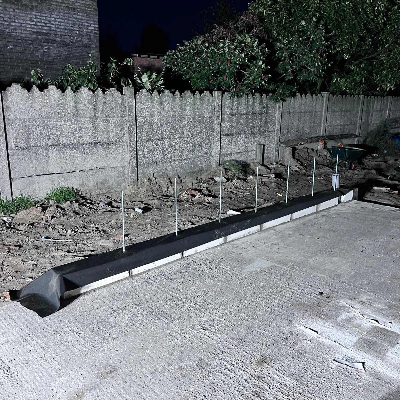 Een tuinperk dat wordt klaargemaakt met afboording en palen, naast een betonnen trottoir en gescheiden van een grijze betonnen schutting door een laagje aarde en kleine plantjes.