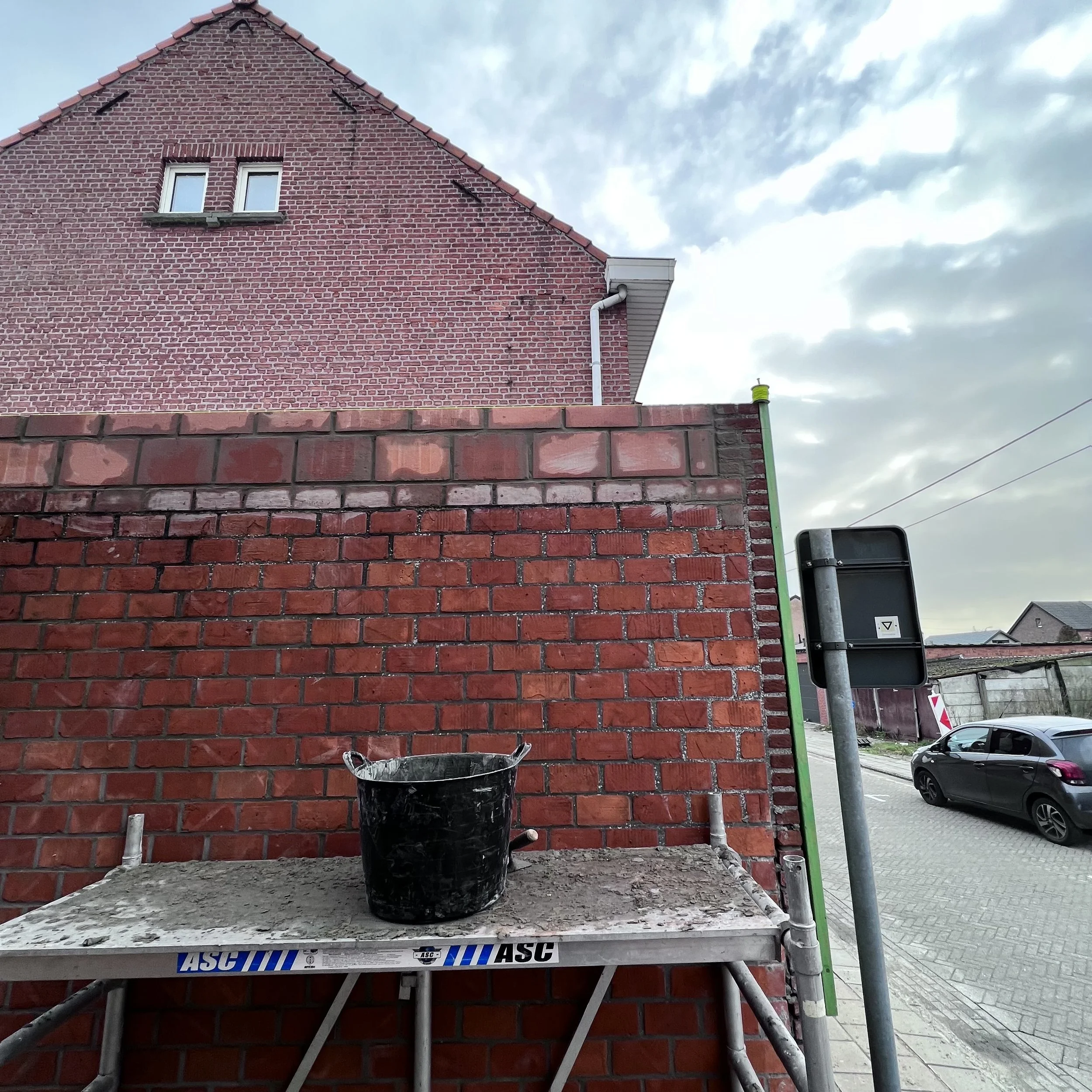 Een bouwplaats met een rode bakstenen muur, een zwarte emmer op een stelling en een straat met geparkeerde auto's, onder een bewolkte hemel.