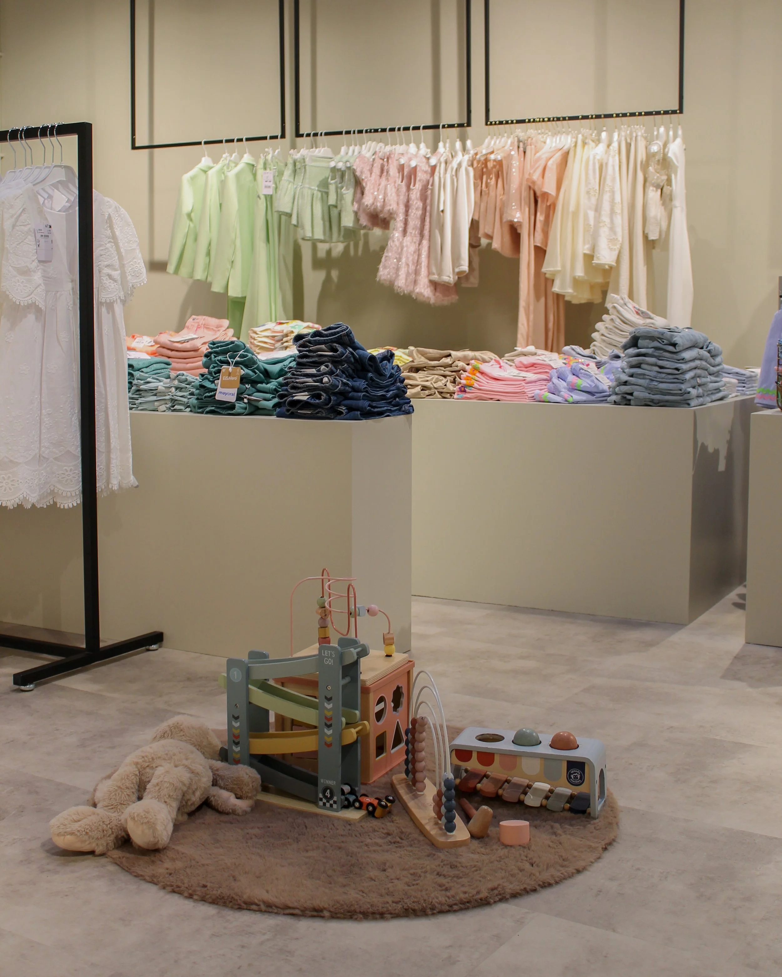 Kinderkleding staat uitgestald op rekken en tafels, en er is een speelhoek op een bruin tapijt met knuffels en houten speelgoed.
