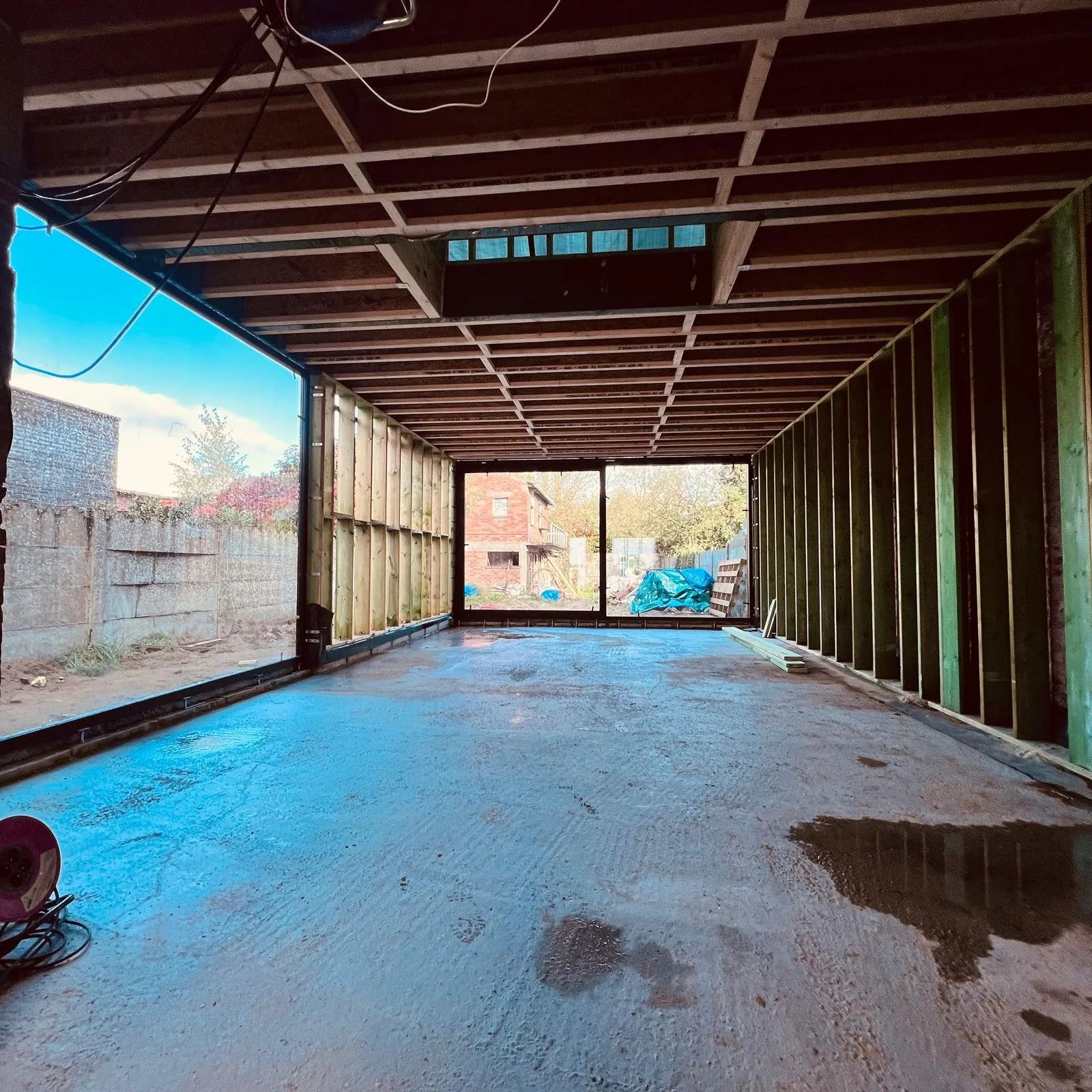 Interieur van een gebouw in aanbouw met onafgewerkte muren, een betonnen vloer en grote ramen die uitzicht bieden op de buitenomgeving met bomen en bouwmaterialen.