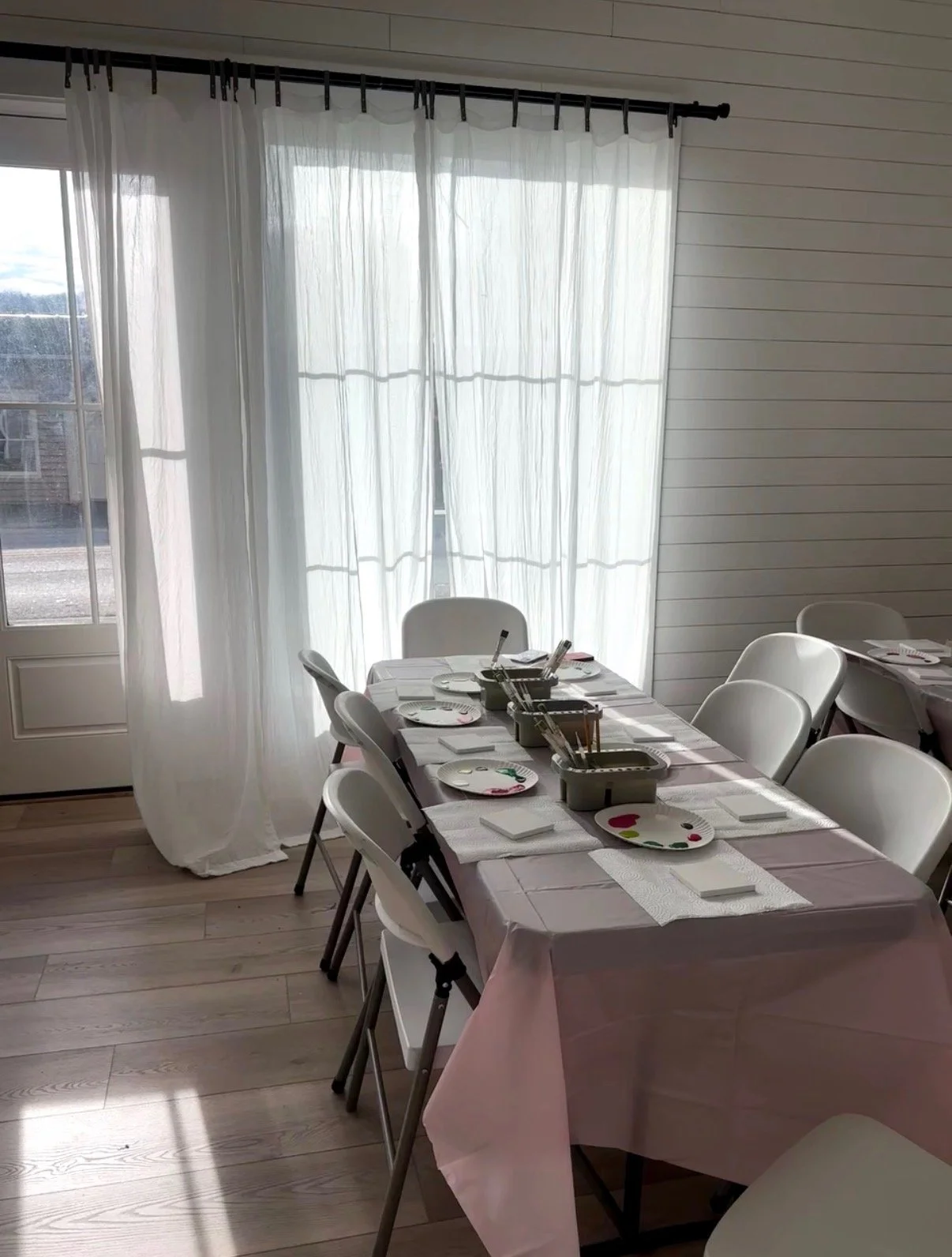 A dining room with a table set for a meal. The table has a pink tablecloth, white plates with colorful hearts, napkins, and utensils. There are six white chairs around the table. Sheer white curtains cover large windows letting in natural light. The floor is light wood, and the walls are white with horizontal paneling.