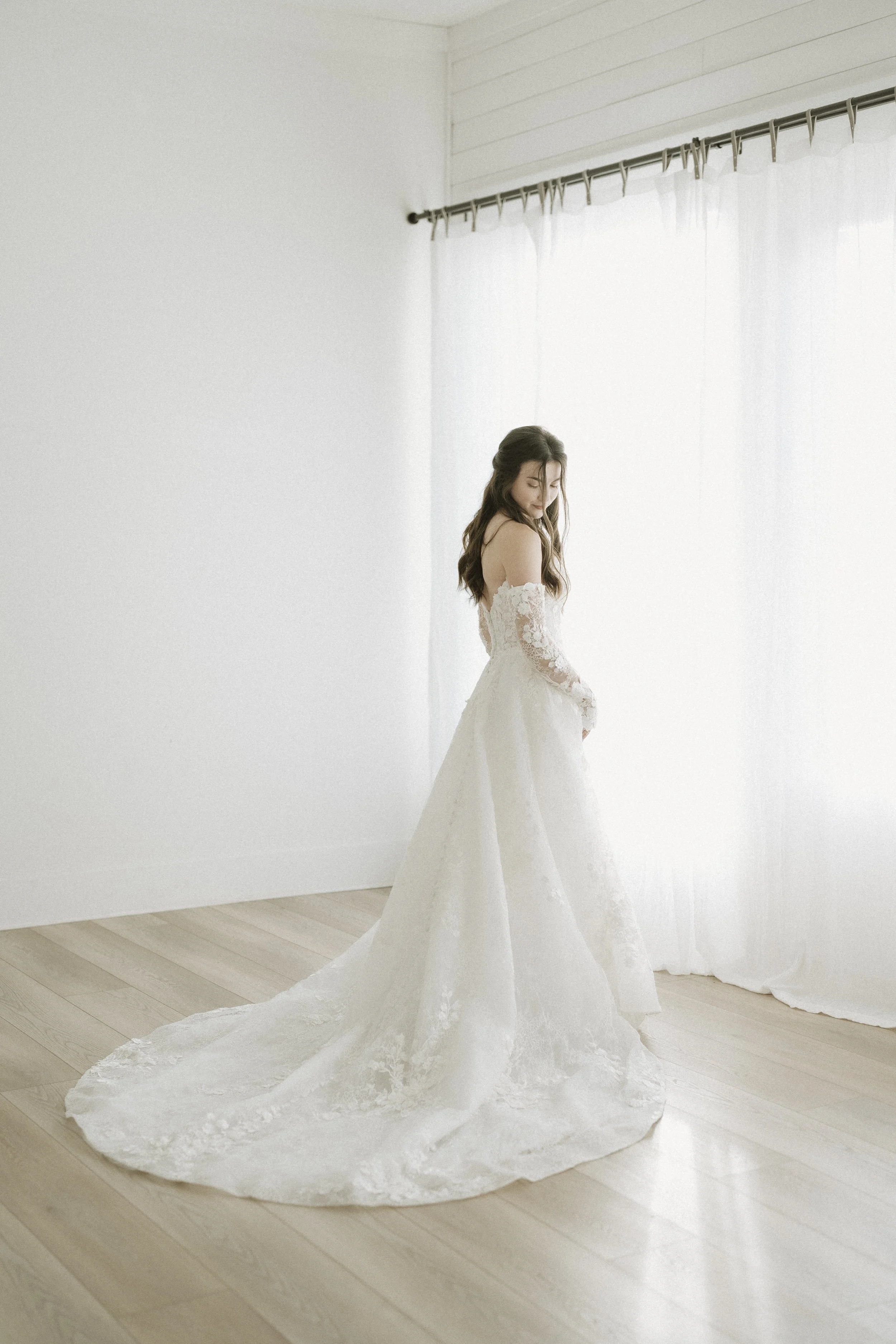 A woman in a white wedding dress standing on a wooden floor near white curtains, looking down.