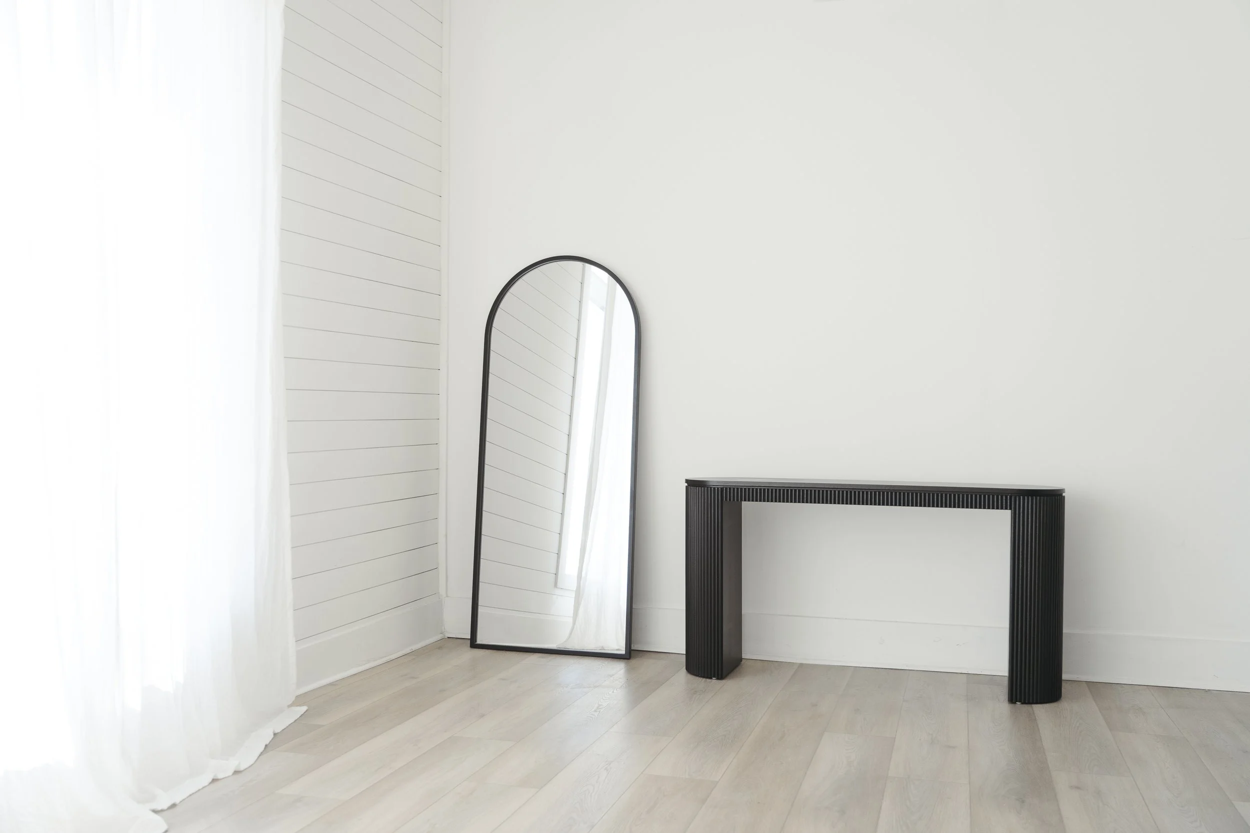 Minimalist interior scene with a black console table, a tall free-standing mirror with a black frame, white shiplap wall, and sheer white curtains in a bright room.